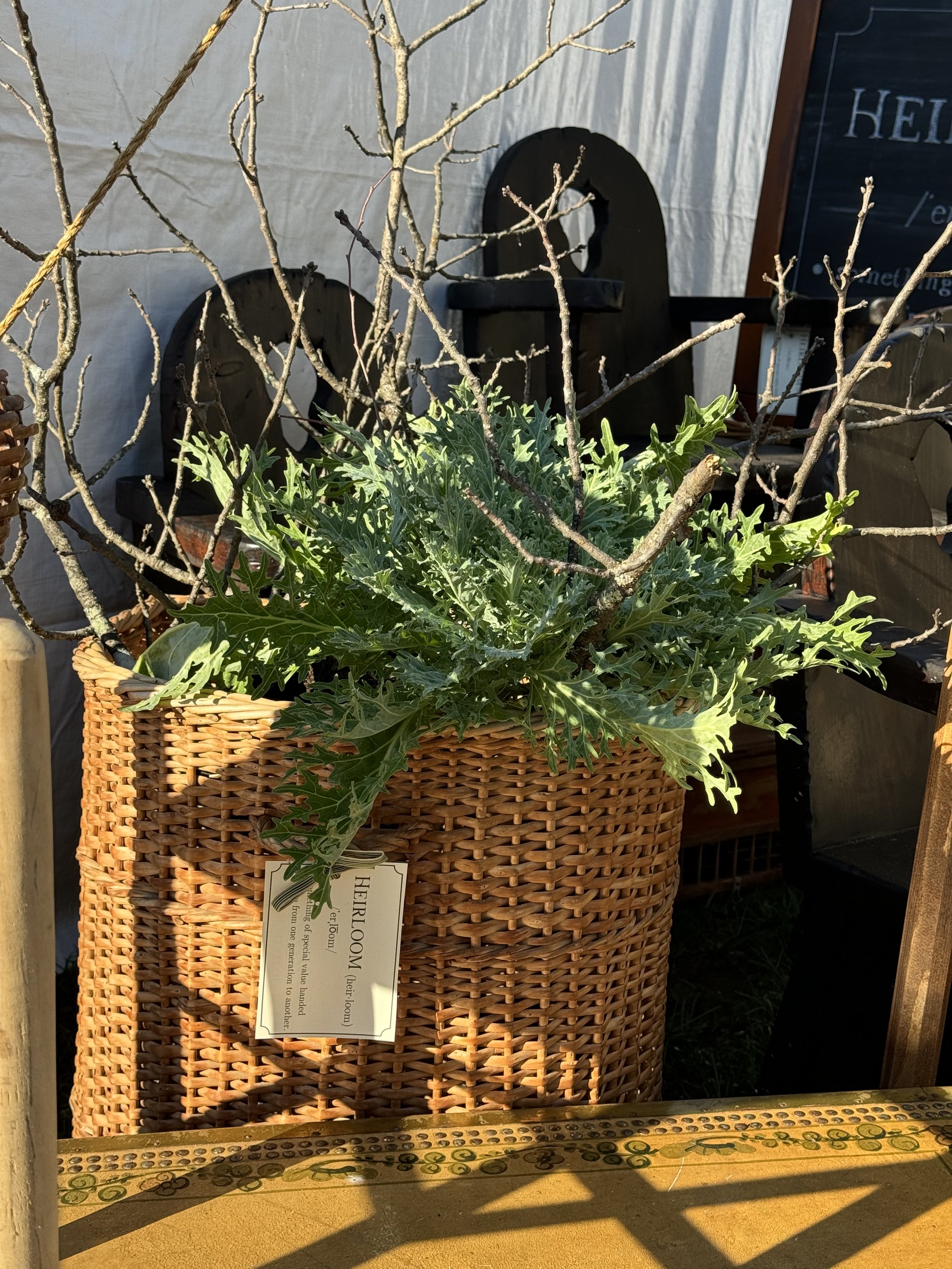 A wicker basket containing green leafy plants and bare branches, with a tag labeled "HEIRLOOM." It is situated in front of a sign with partial text and other objects in the background.