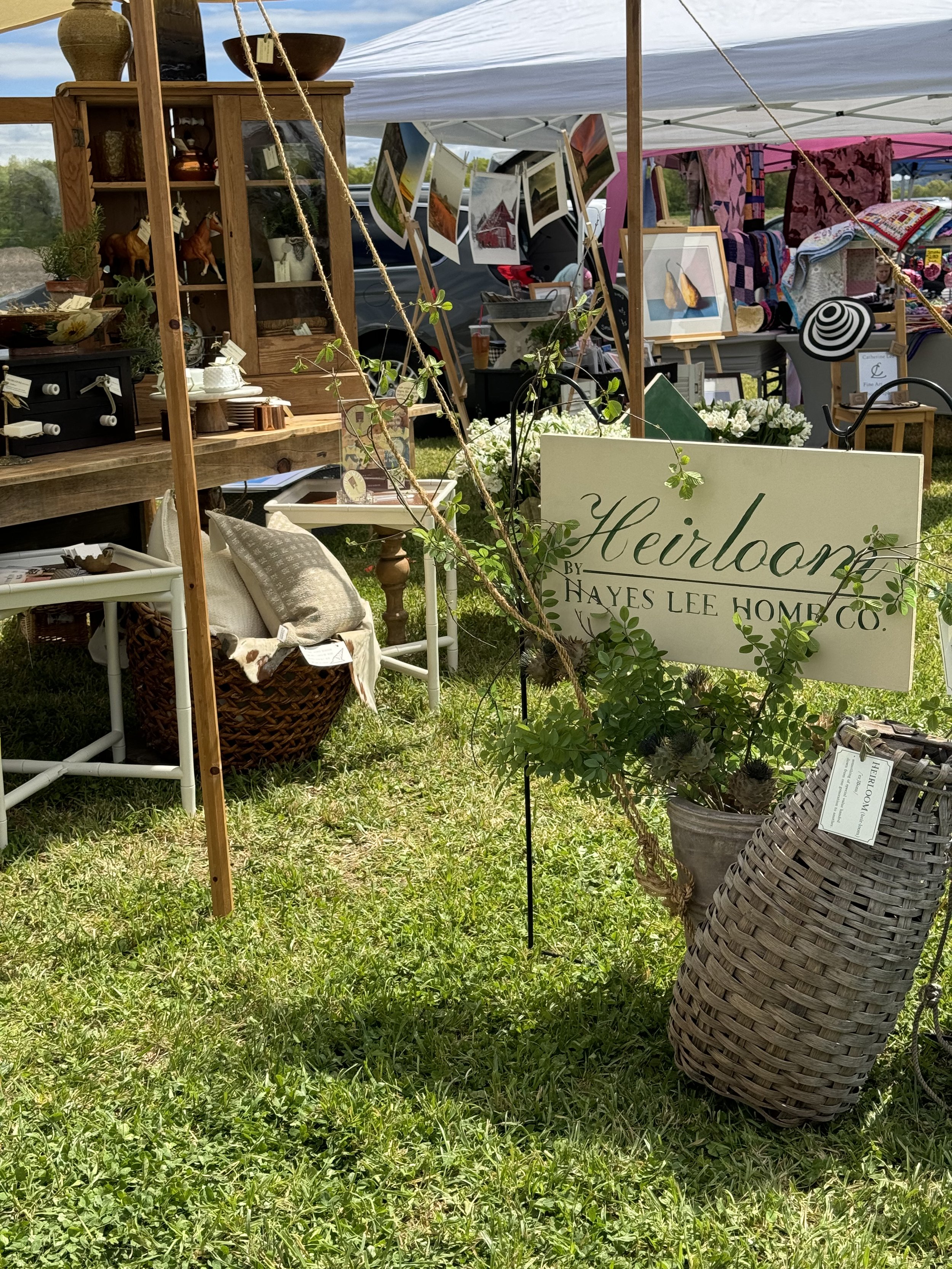Outdoor market stall with vintage and handmade items, wooden furniture, decorative signs, plant baskets, and art prints under a canopy. "Heirloom by Hayes Lee Home Co." sign in foreground.