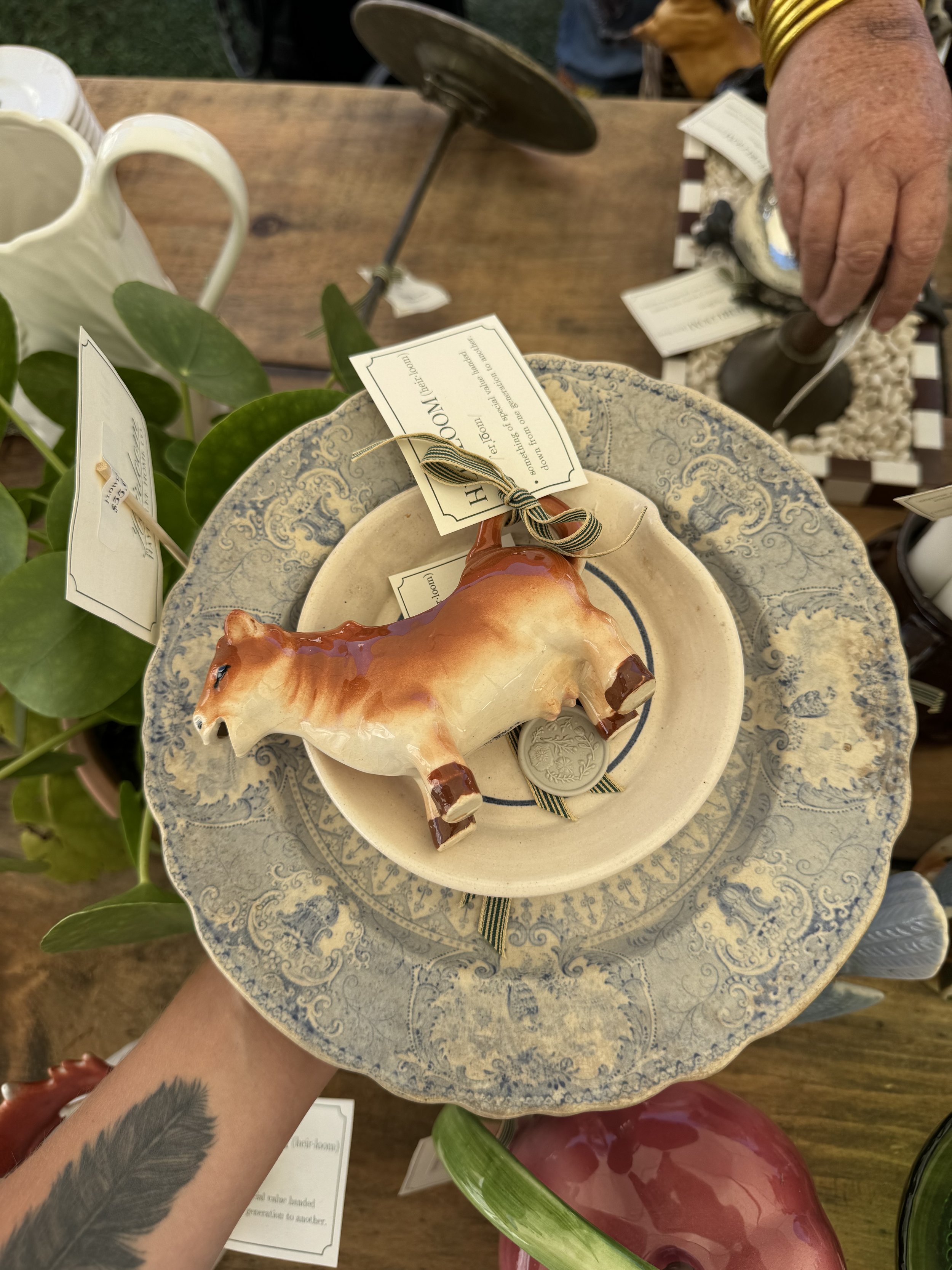 A decorative ceramic cow figurine on two stacked vintage blue patterned plates with vintage-style labels and a plant in the background.