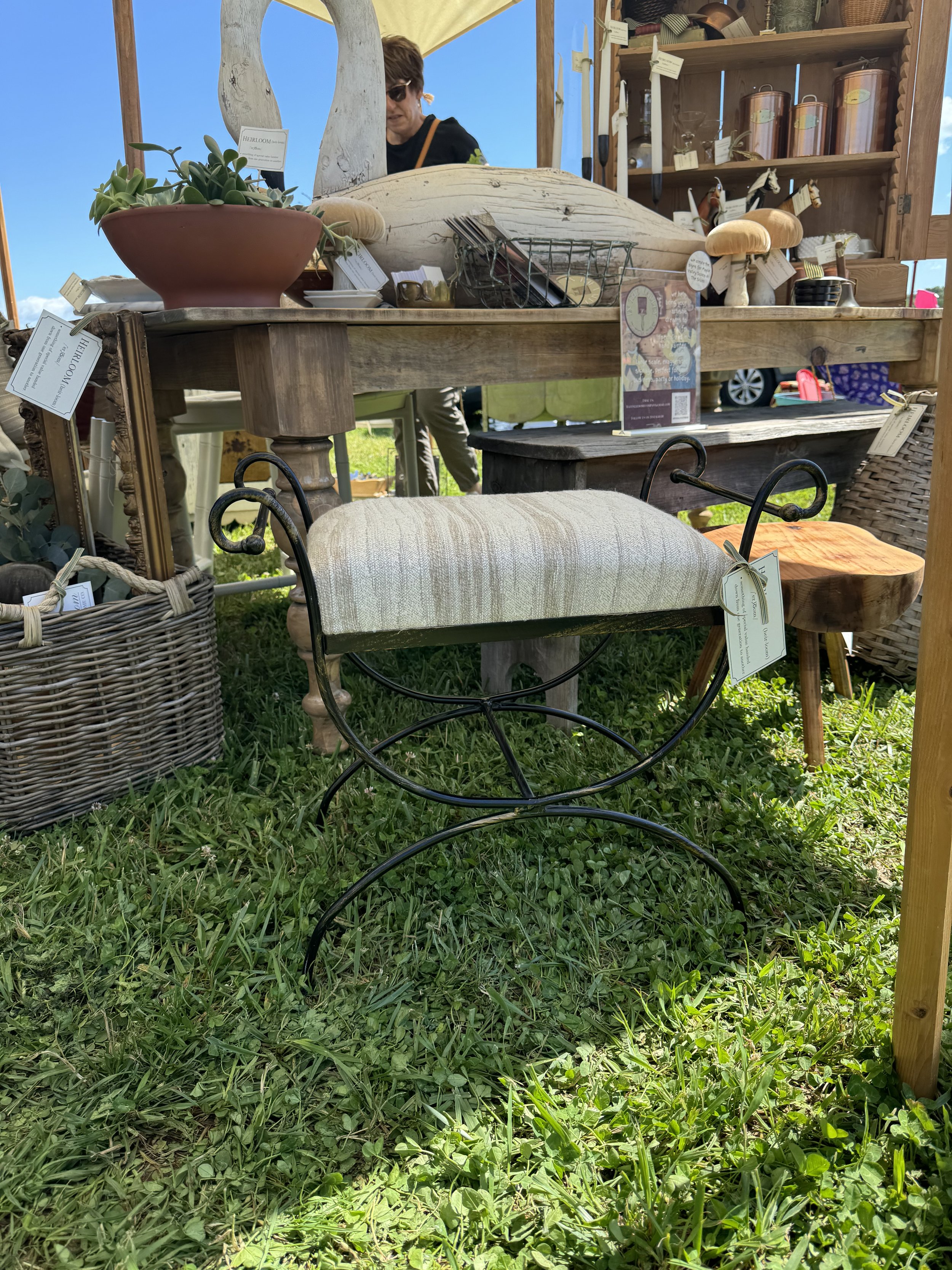 Outdoor flea market booth with vintage furniture and decor, including a metal and upholstered stool, potted plants, wooden shelves with items, and wicker basket on grass.