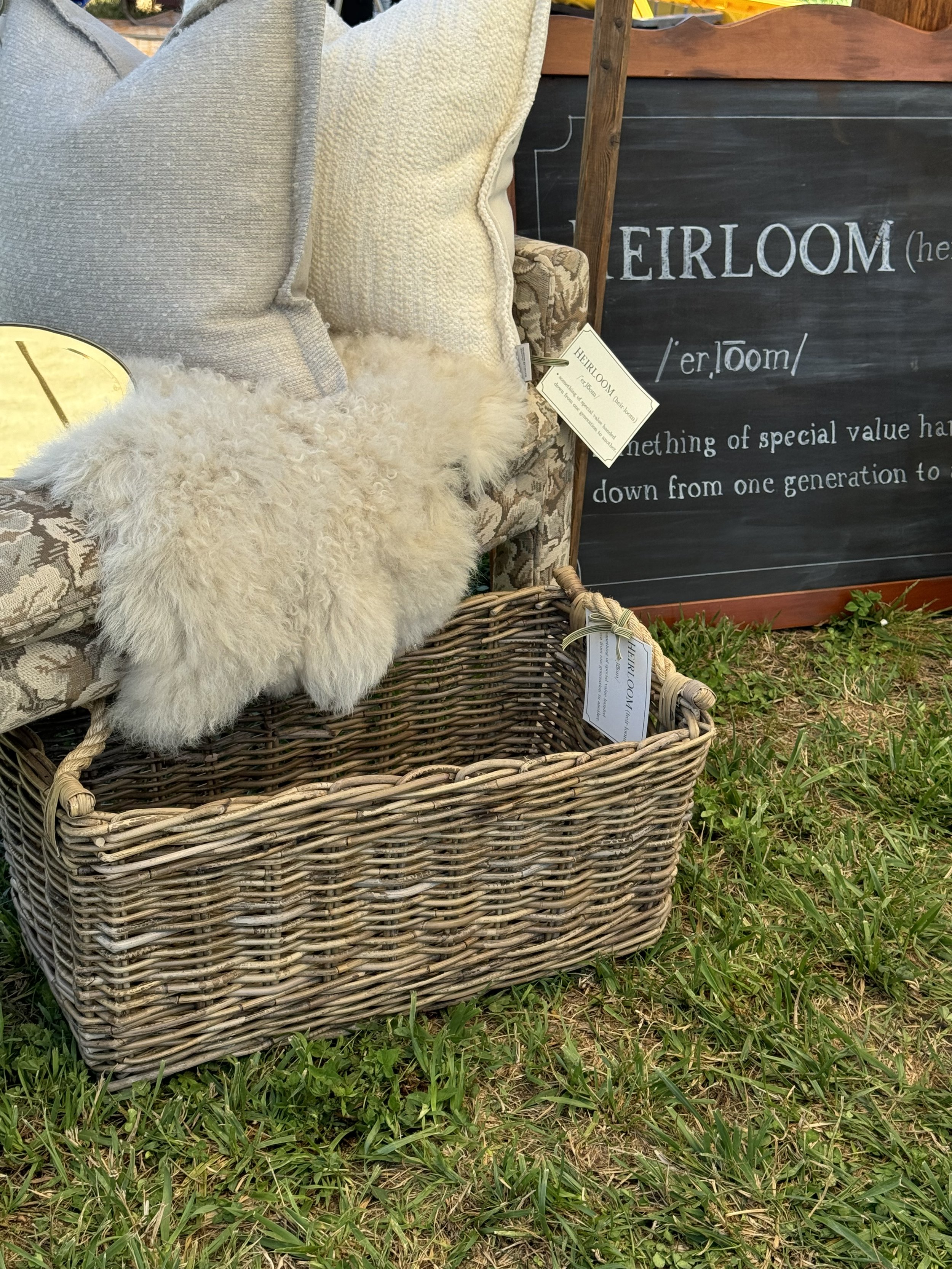 Wicker basket on grass with pillows and sheepskin atop, near chalkboard with 'Heirloom' definition.