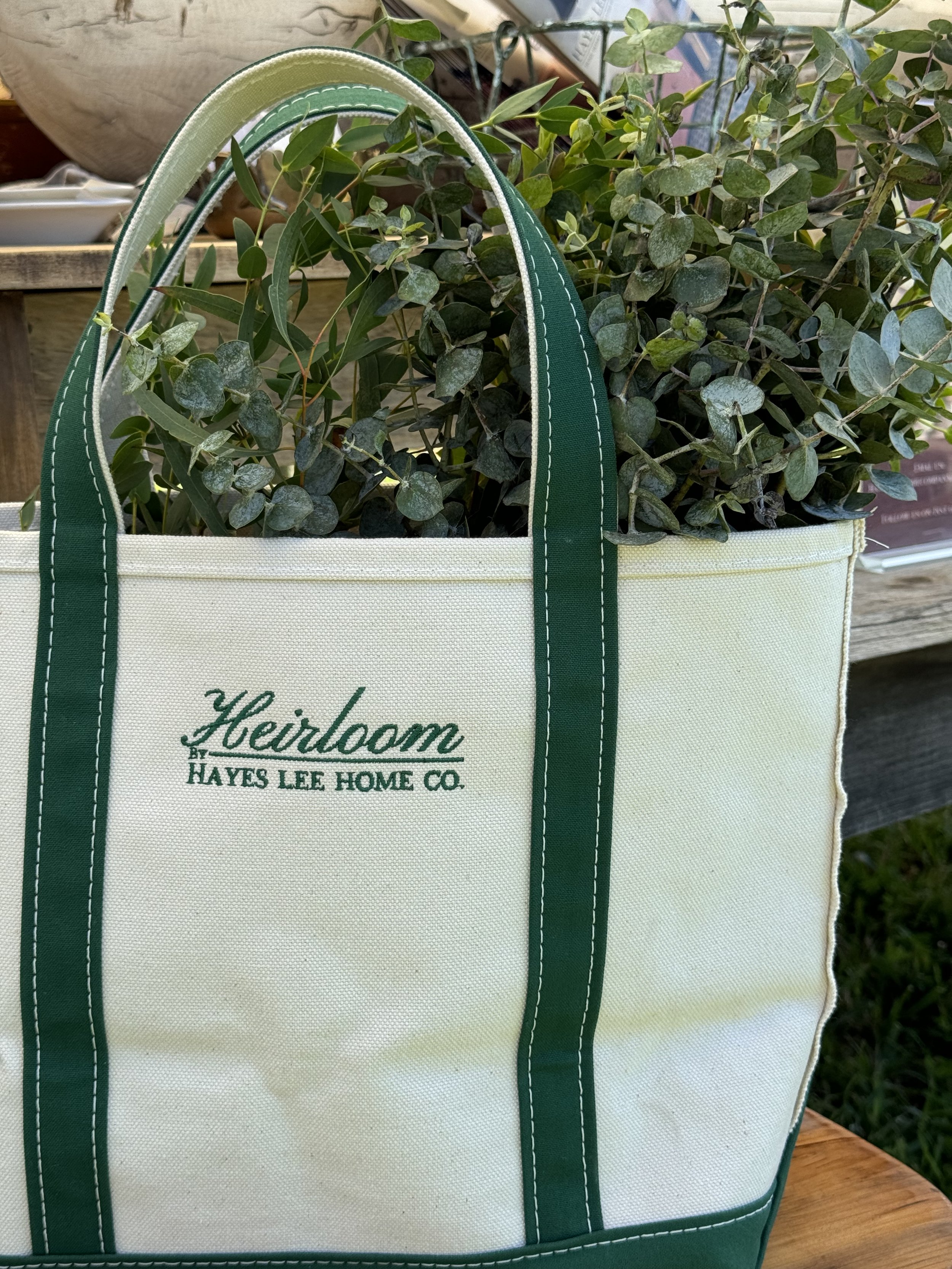 Canvas tote bag with green handles containing eucalyptus branches, labeled "Heirloom by Hayes Lee Home Co."