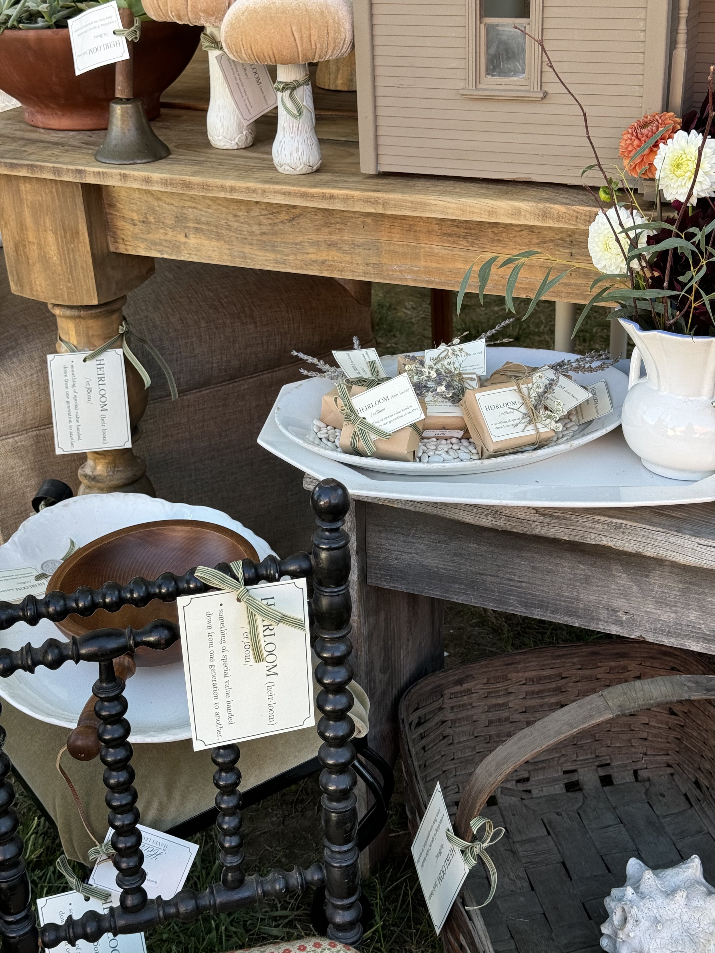Display of vintage items including a wooden table, decorative mushrooms, a miniature house, white ceramic pitcher with flowers, wrapped soap bars, a black spindle chair, and a woven basket.