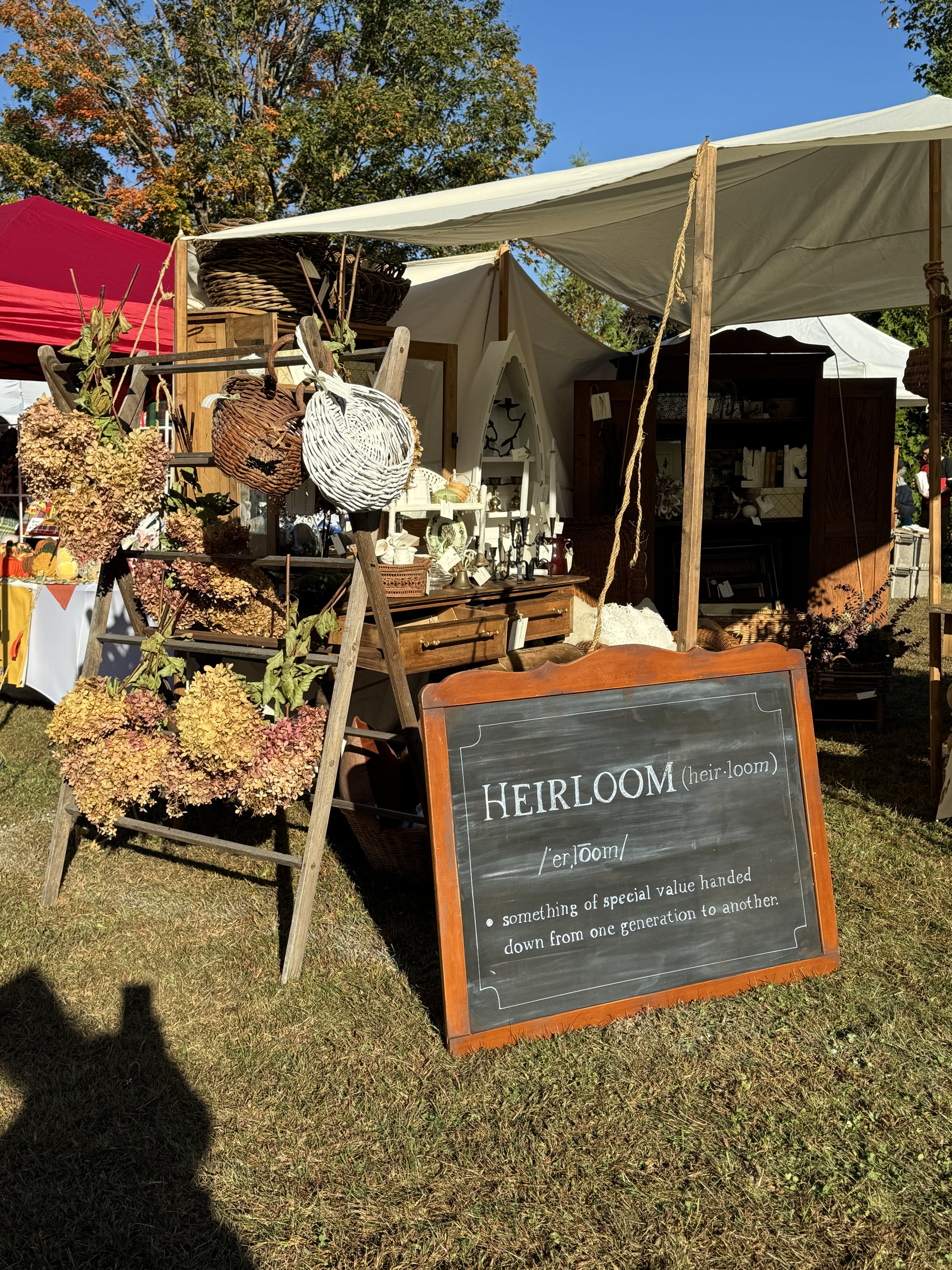 Outdoor market stall with a vintage theme, featuring dried flowers, wicker baskets, and a chalkboard sign displaying the definition of "heirloom." Trees with autumn foliage are visible in the background.