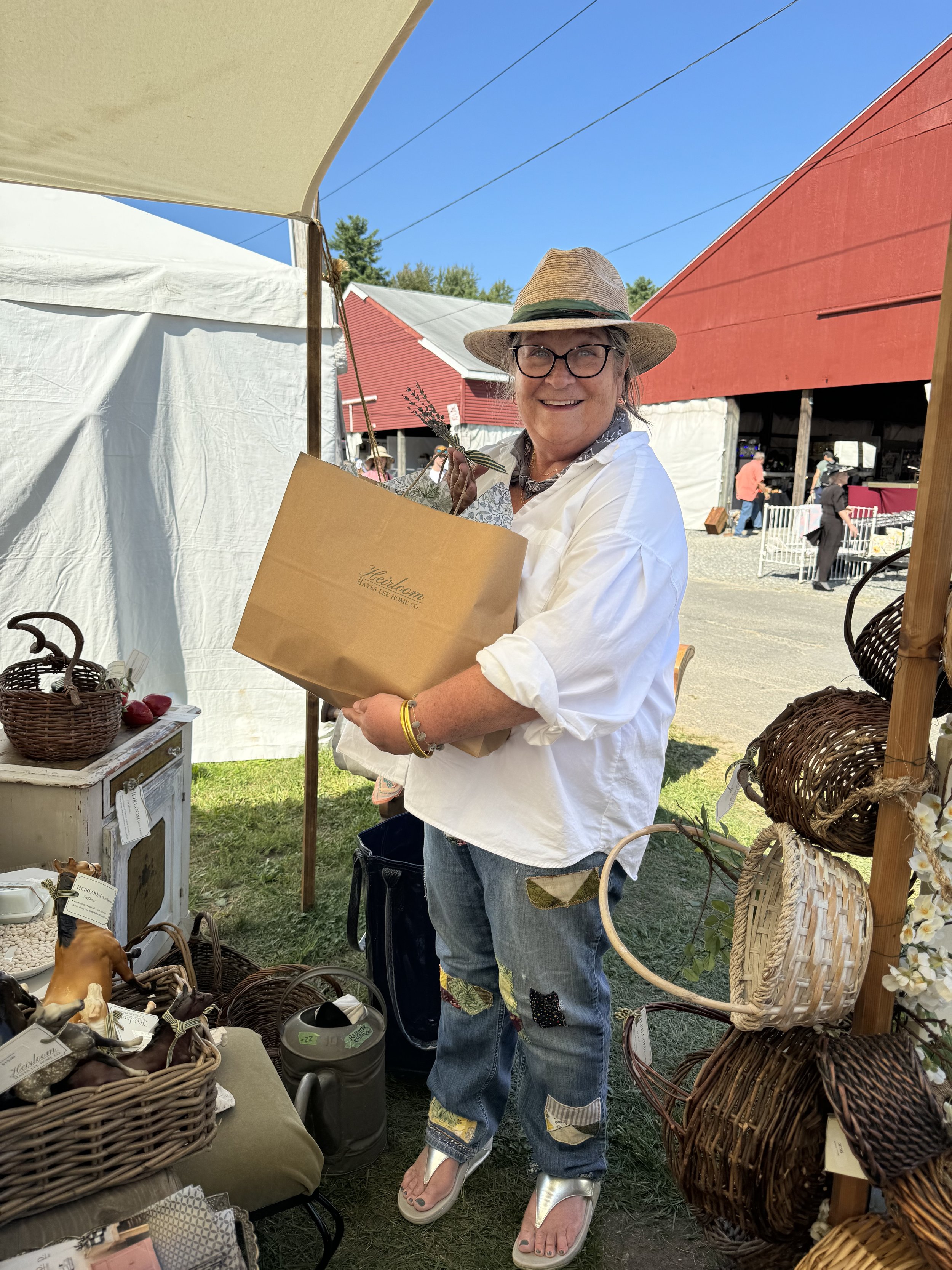 Person smiling while holding a brown shopping bag at an outdoor market with wicker baskets and goods on display.