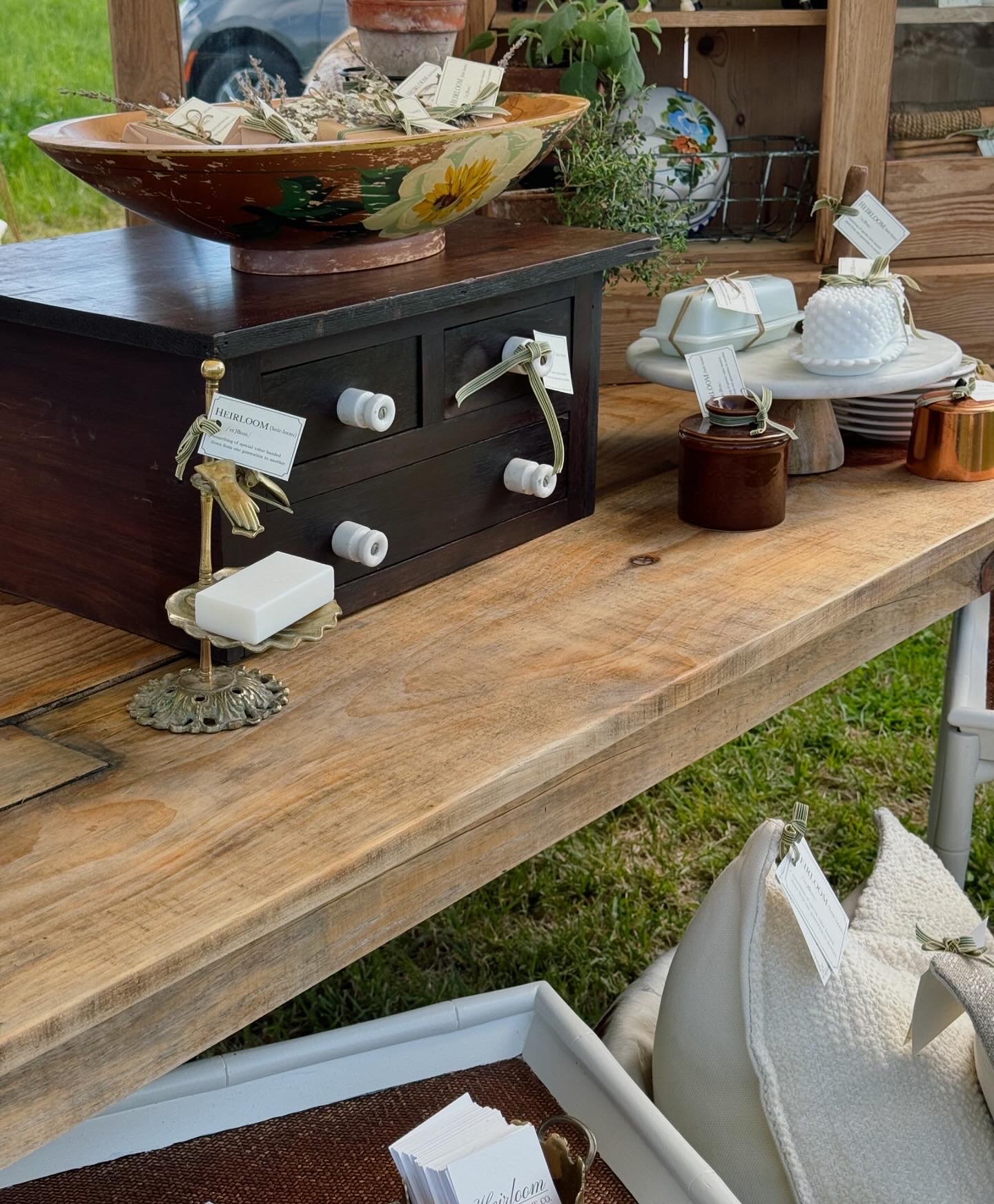 Wooden table displaying various home decor items including a decorative bowl, black drawer unit, white ceramic pieces, and pillows with tags in an outdoor setting.
