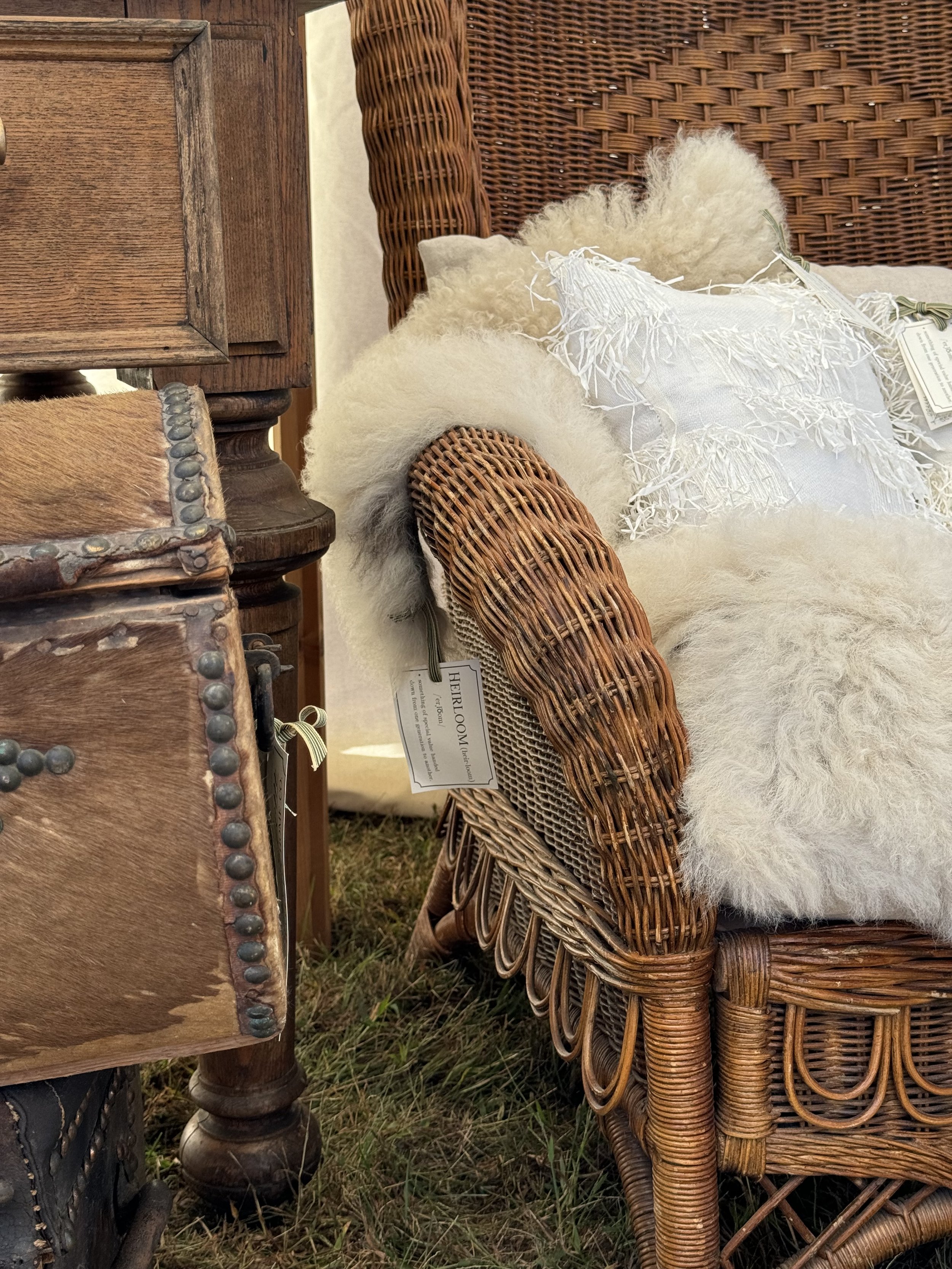 Vintage wicker chair with white throw and decorative pillows, next to a wooden table and a cowhide trunk, displayed on grass.