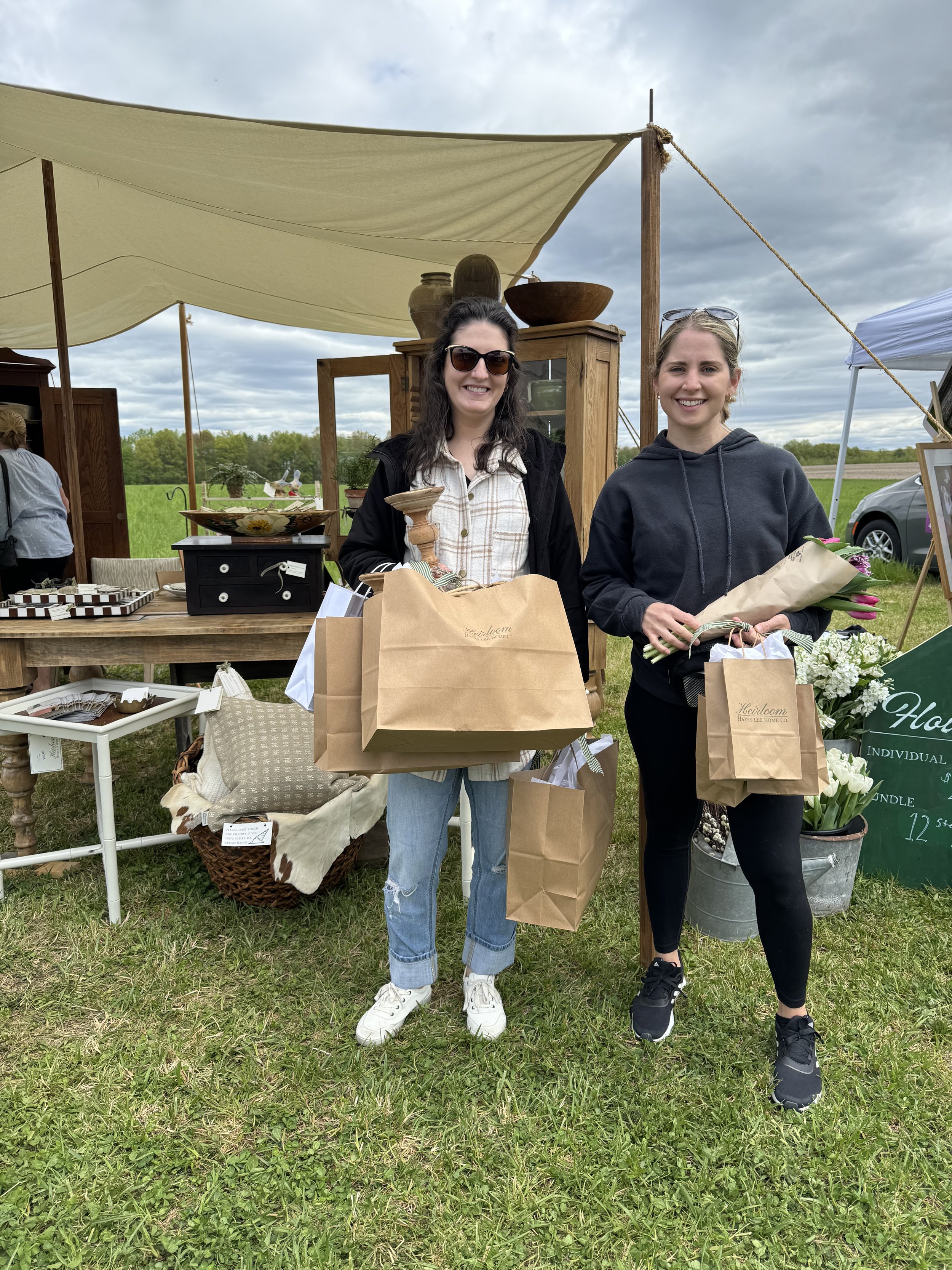 Two women holding paper shopping bags at an outdoor market, standing on grass in front of a booth with various items displayed. One woman holds flowers wrapped in paper.