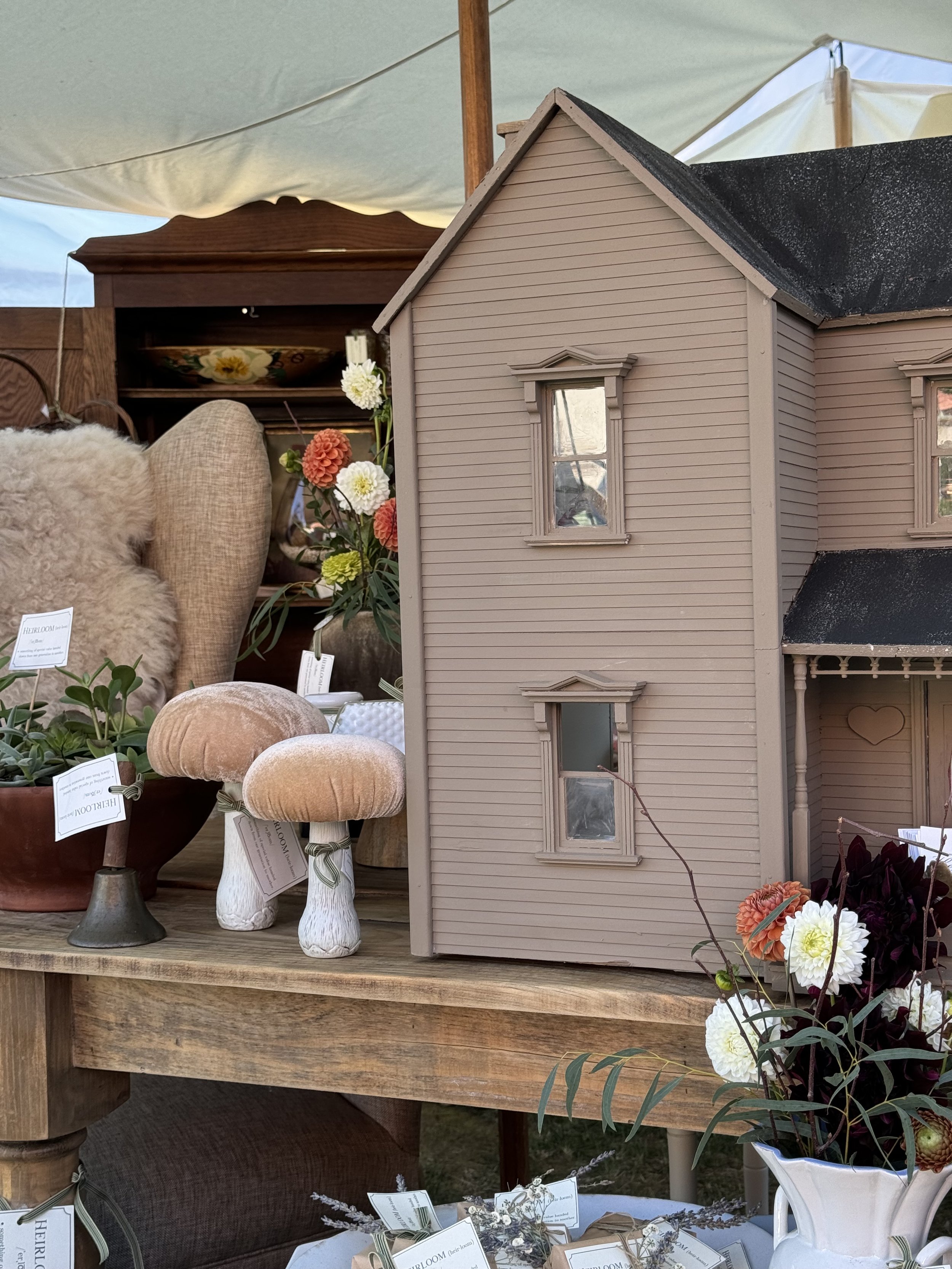 A vintage dollhouse with a brown exterior is displayed on a wooden table, surrounded by decorative items such as plush mushrooms and vases of flowers. The background includes fluffy textiles and rustic furniture pieces.