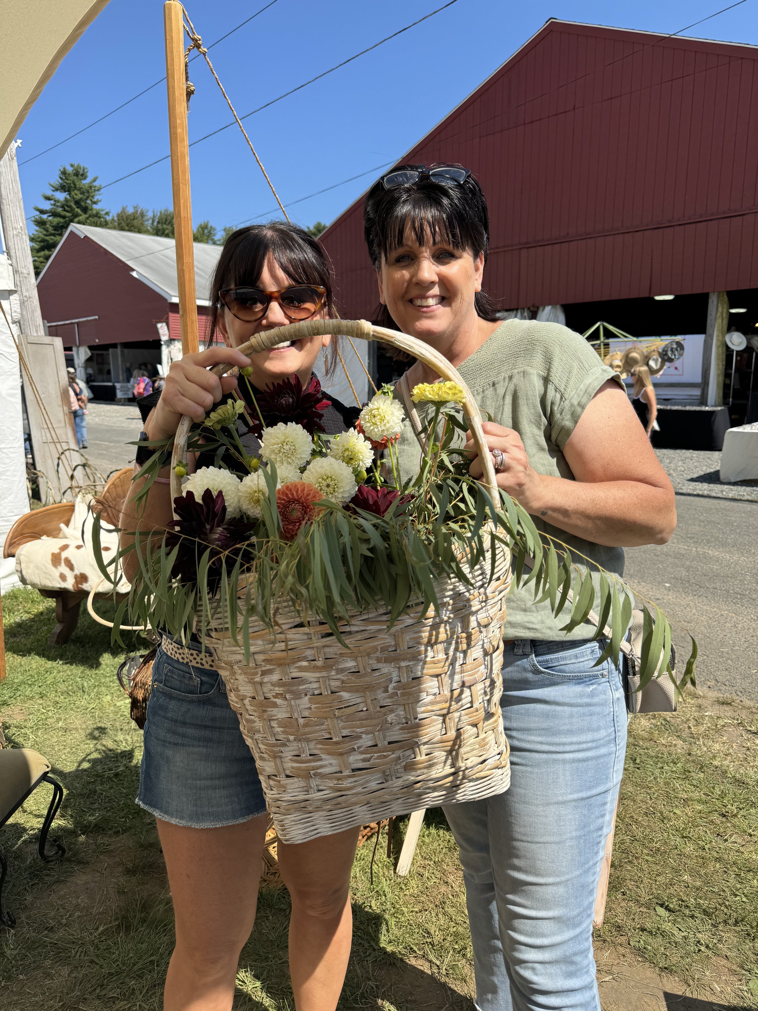 Two women smiling and holding a large basket with colorful flowers outdoors at a fair or festival, with a red barn in the background.