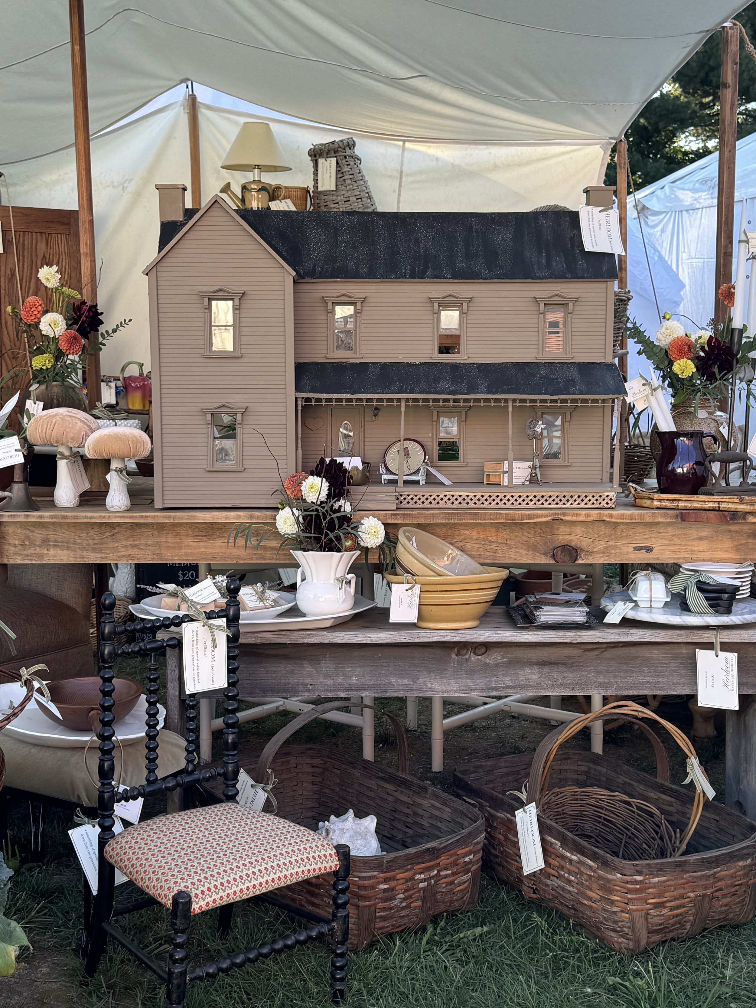 Antique market booth with a wooden dollhouse, various vases with flowers, wicker baskets, pottery bowls, and a small antique chair on display under a white tent canopy.