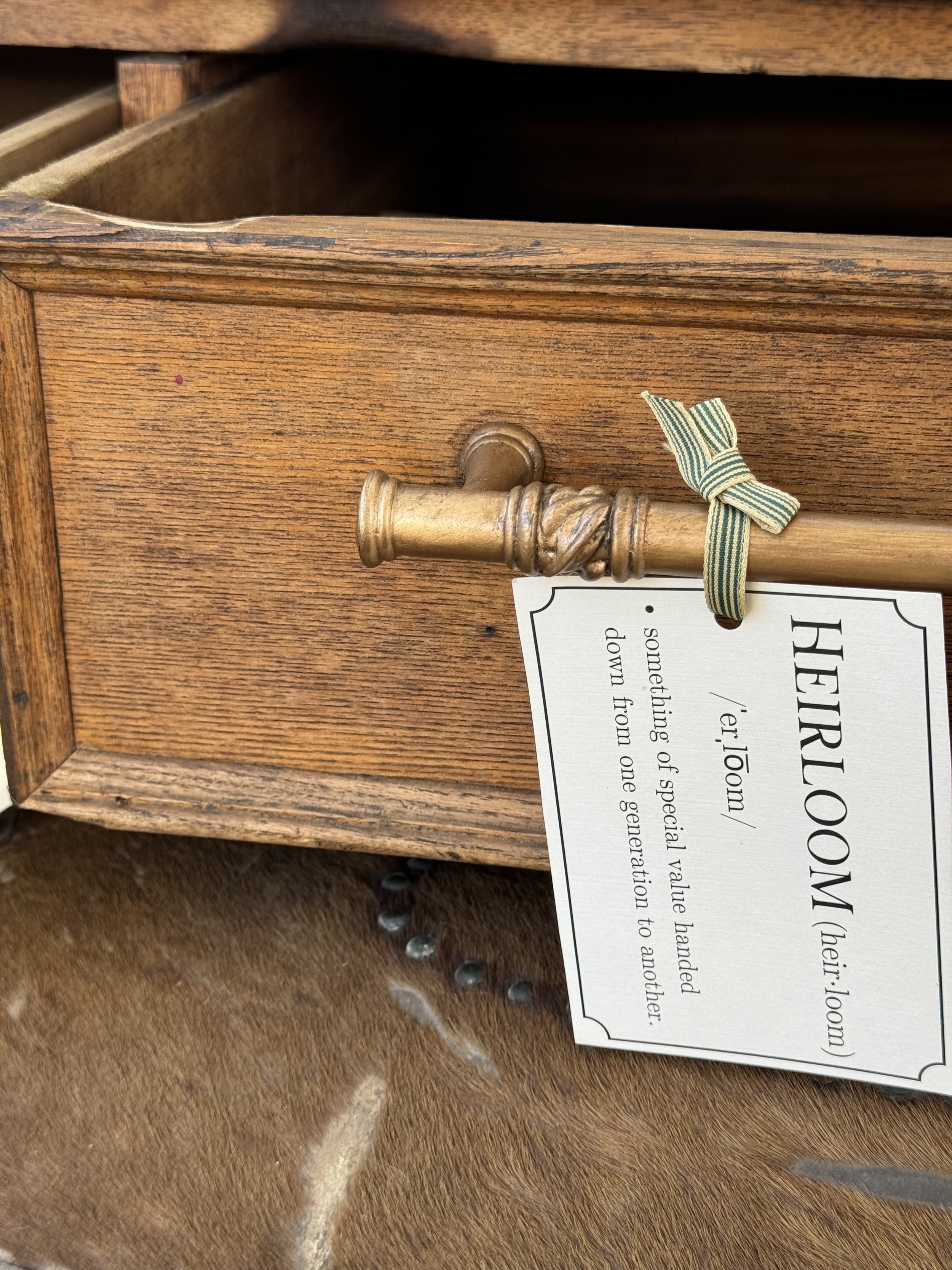 Close-up of an open wooden drawer with a brass handle, featuring a green ribbon and a tag labeled 'HEIRLOOM' with its definition.