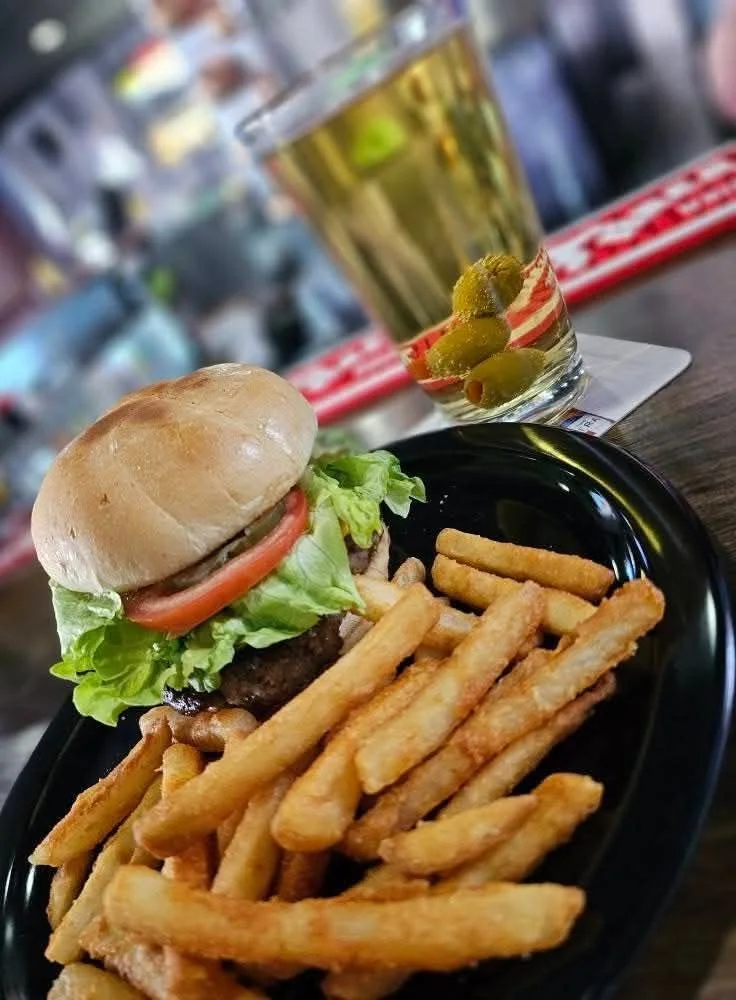 A cheeseburger with lettuce and tomato on a black plate with French fries, next to a glass of beer garnished with olives on a bar counter.