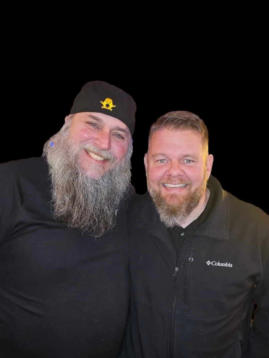 Two men standing together, both smiling, with beards. The man on the left wears a beanie with a yellow logo, while the man on the right wears a Columbia jacket. The background is black.
