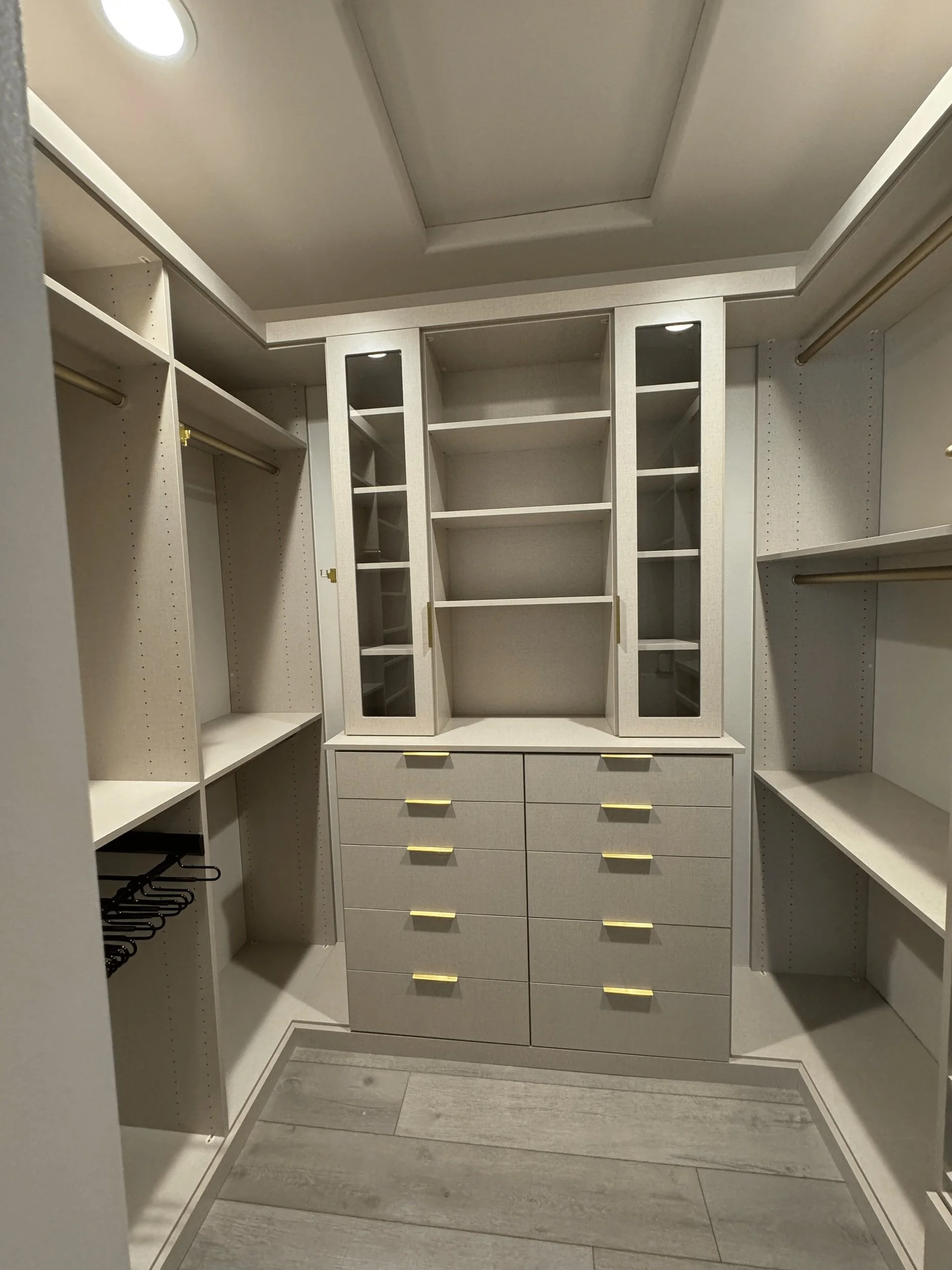 Modern walk-in closet with shelving, drawers, and hanging rods, featuring beige cabinets and gold handles.