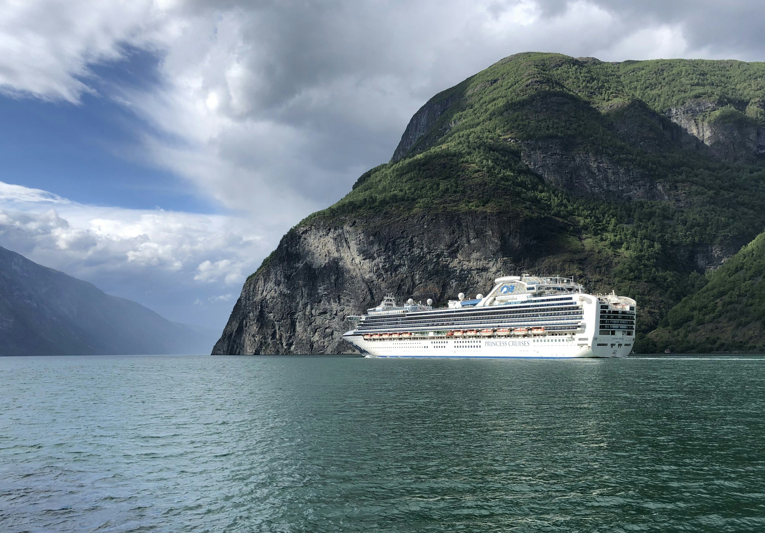 A large white cruise ship sailing on a body of water surrounded by tall green mountains with a partly cloudy sky overhead.