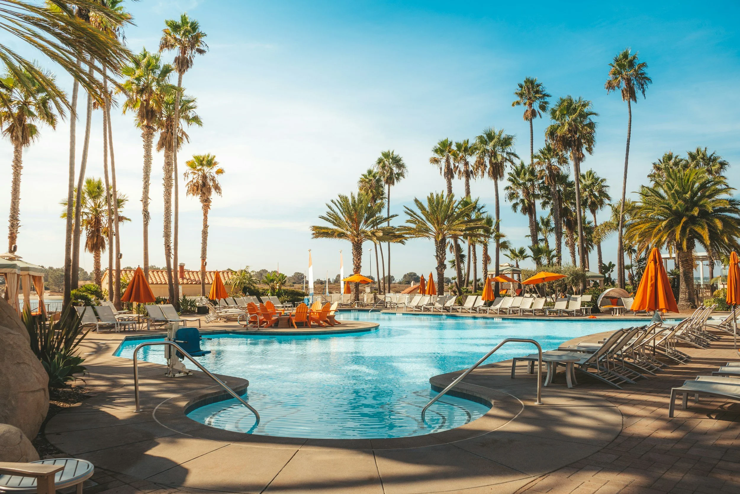 An outdoor swimming pool area with lounge chairs and orange umbrellas, surrounded by tall palm trees under a clear blue sky.