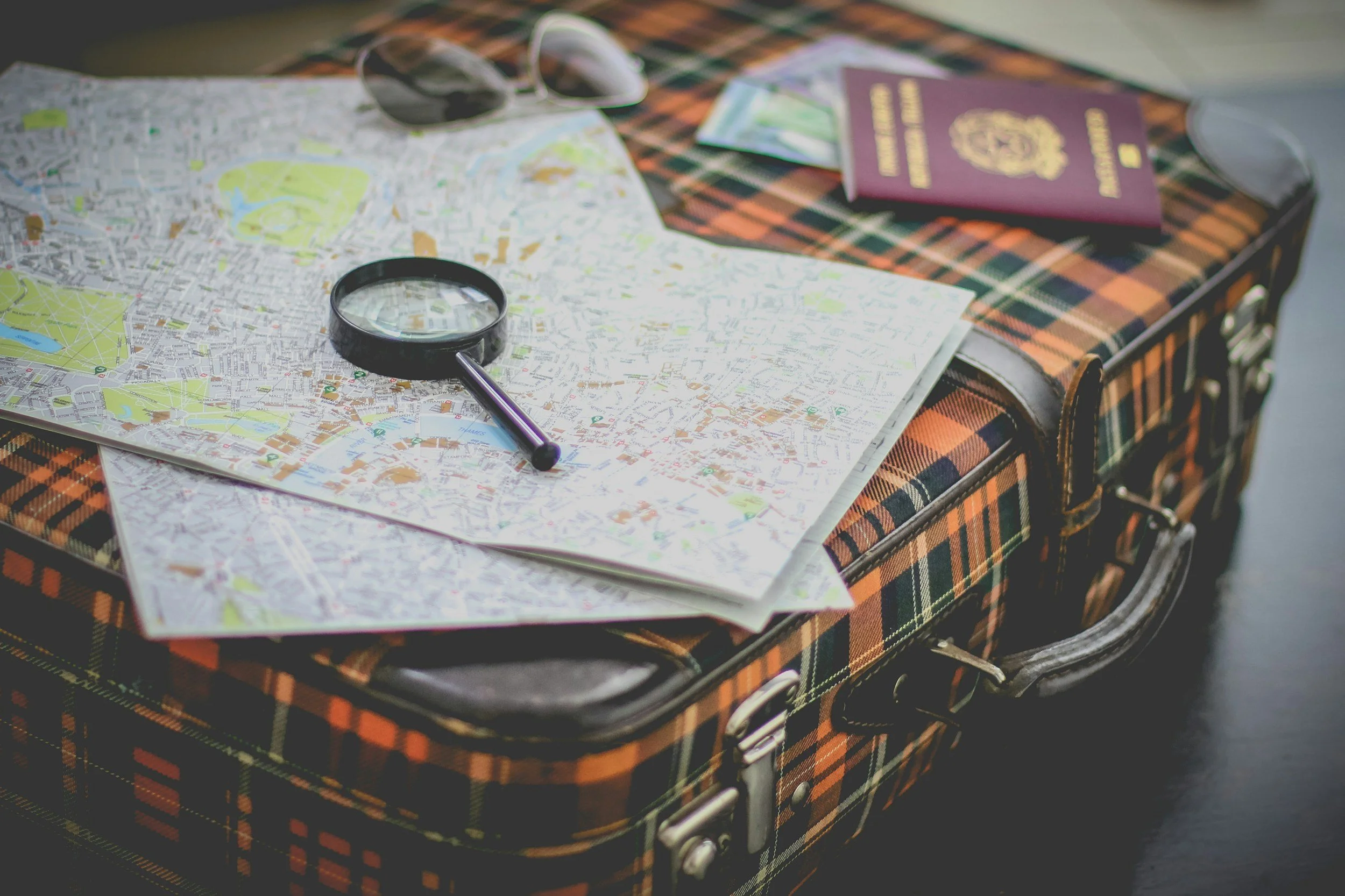 A vintage plaid suitcase on a table with two maps, a magnifying glass, a pair of sunglasses, a passport, and travel tickets.
