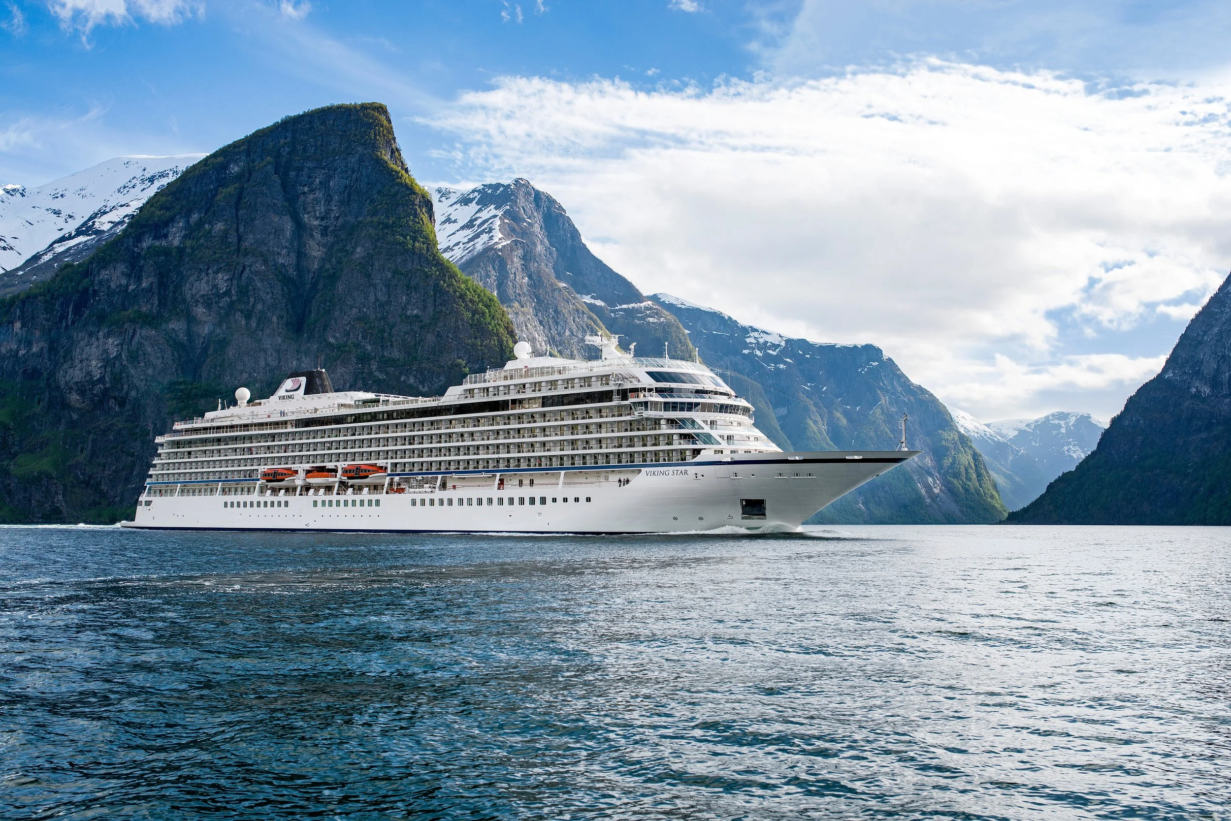 Large white cruise ship sailing through a fjord surrounded by towering, snow-capped mountains and cloudy sky.