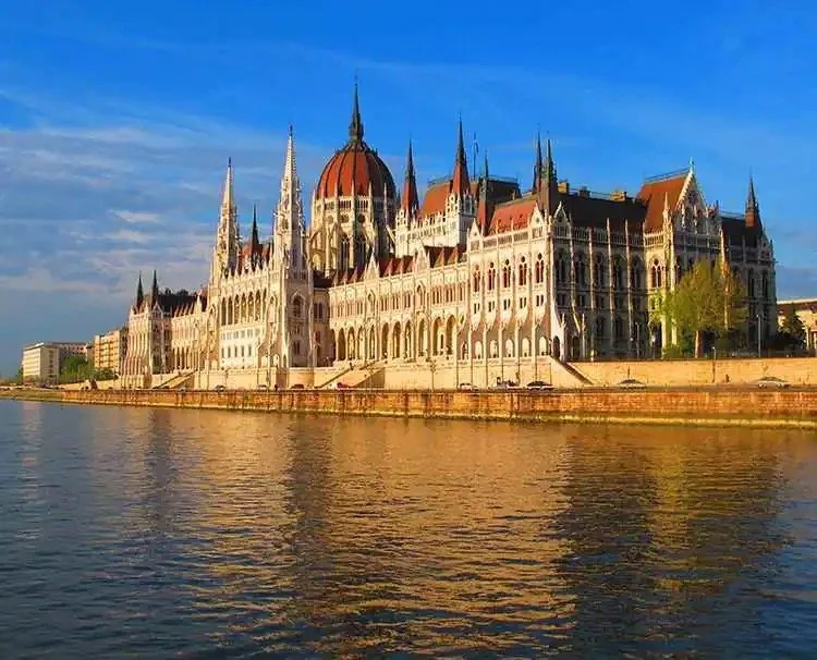The Hungarian Parliament Building along the Danube River during daytime, showcasing its Gothic Revival architecture with spires, a central dome, and detailed facade, under a partly cloudy sky.
