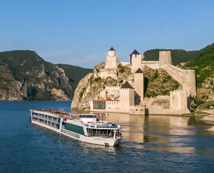 A large river cruise boat sailing on a river with a historic stone castle on a hilltop in the background under a clear blue sky.