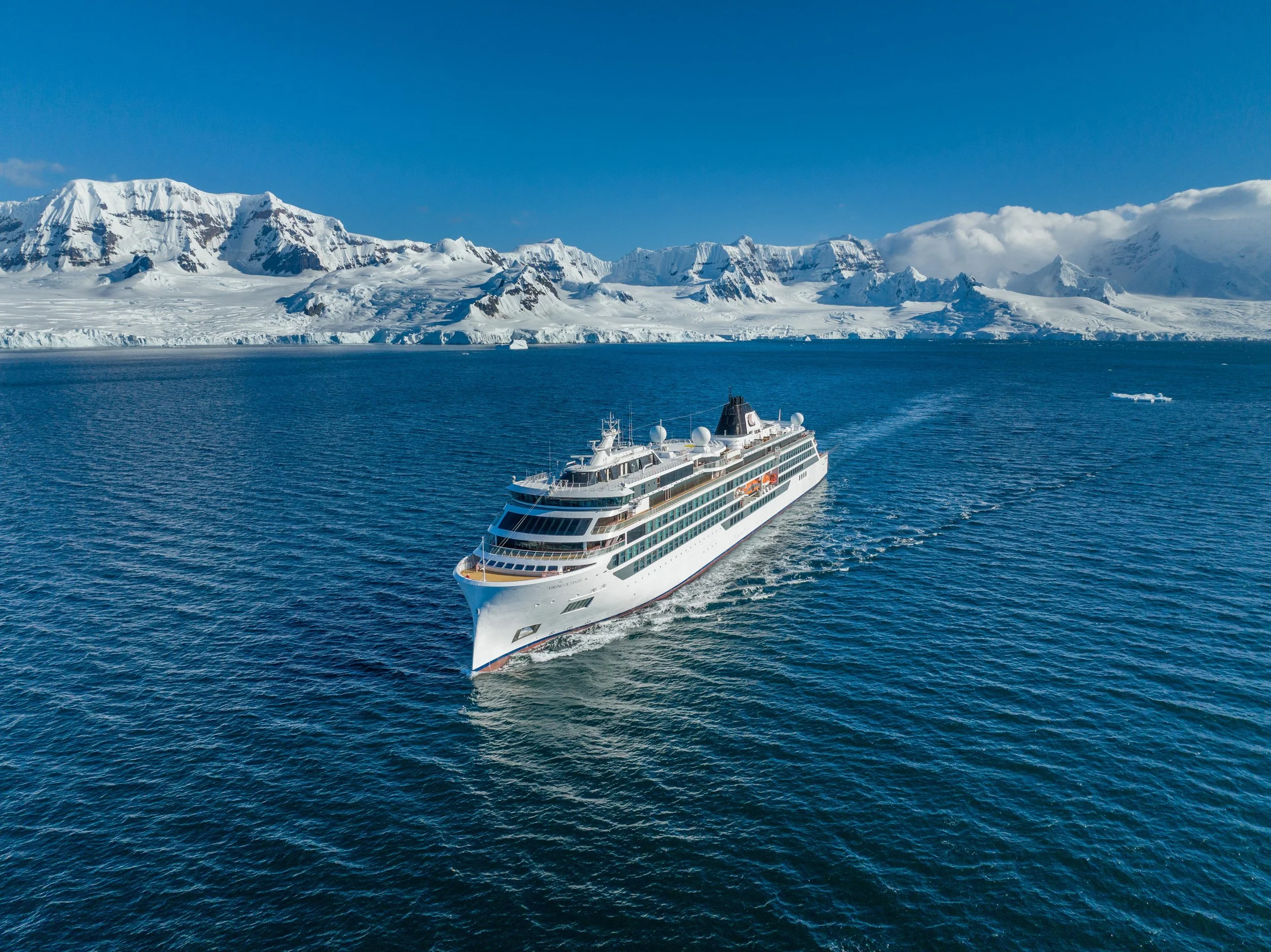 A large white cruise ship sailing in icy blue ocean with snow-covered mountains in the background under a clear blue sky.
