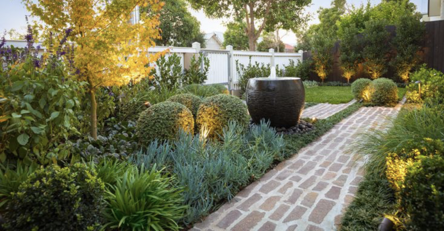 Beautiful garden with a brick pathway, lush greenery, manicured shrubs, a yellow-leaved tree, and a large decorative water feature at a residence in Camberwell, Melbourne.