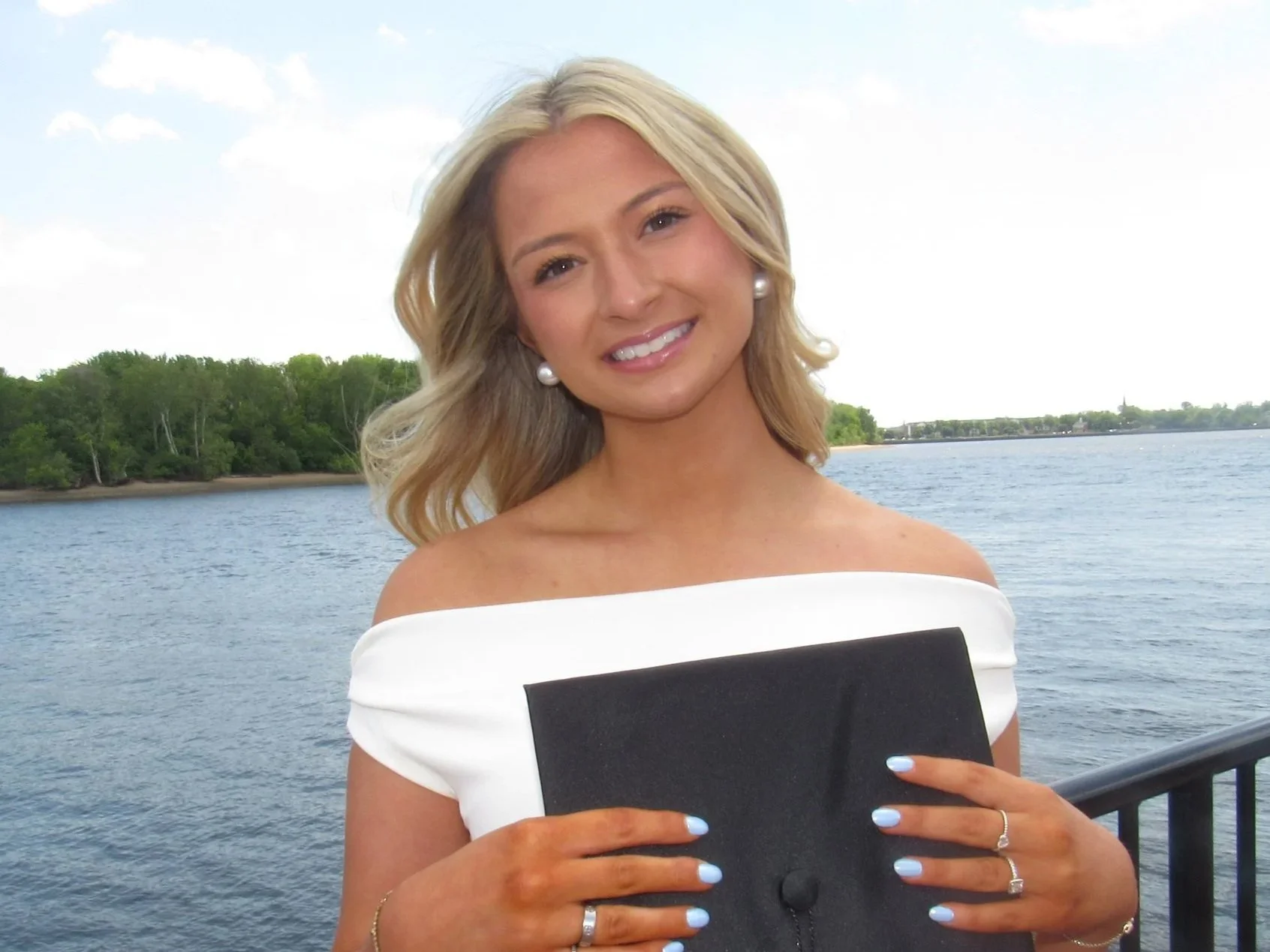 A woman with blonde hair smiling while holding a black gift box in front of her, standing near a body of water with trees in the background.