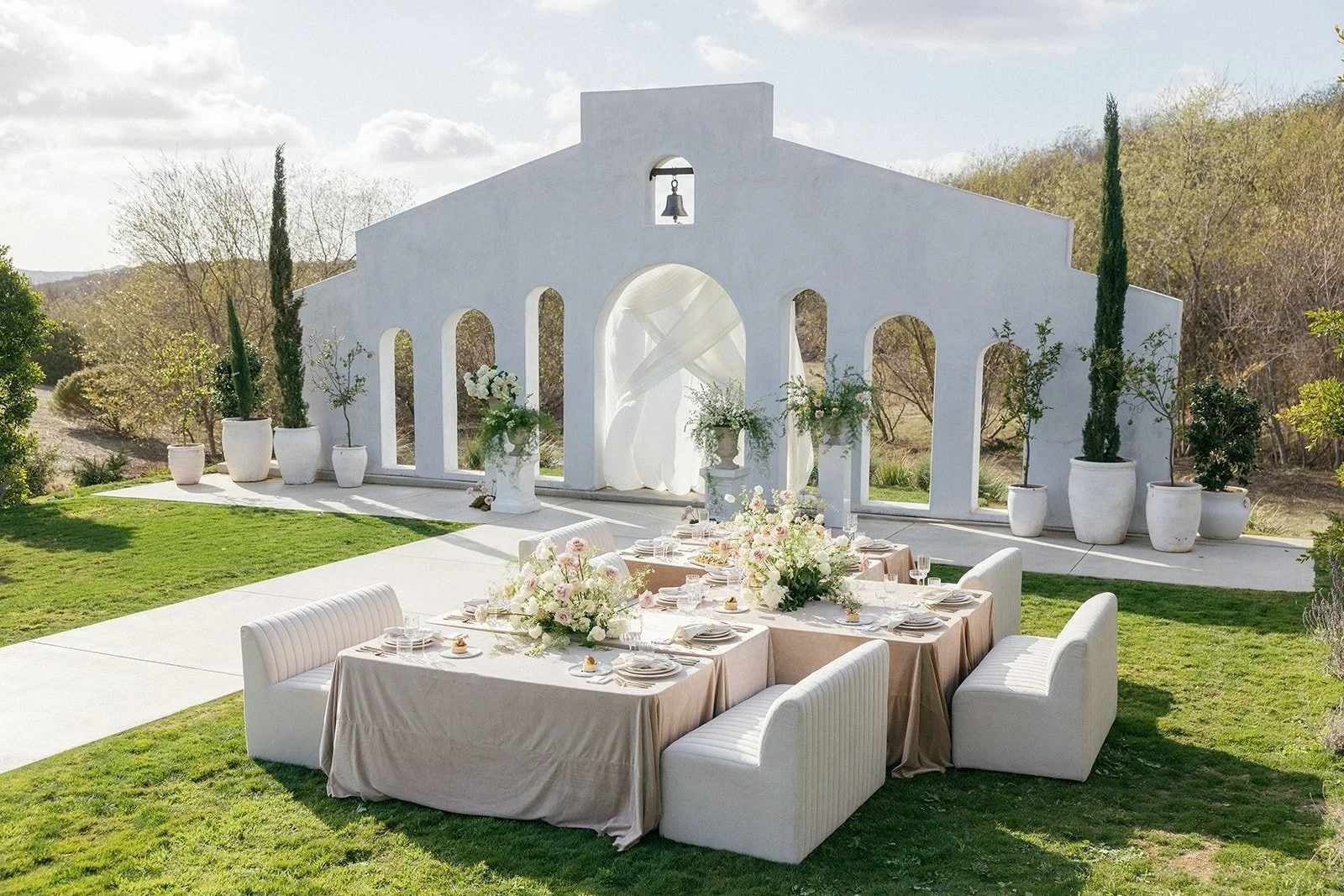 Outdoor wedding reception setup with a white arch structure, elegant tables with beige tablecloths, floral arrangements, and white chairs on a grassy lawn.