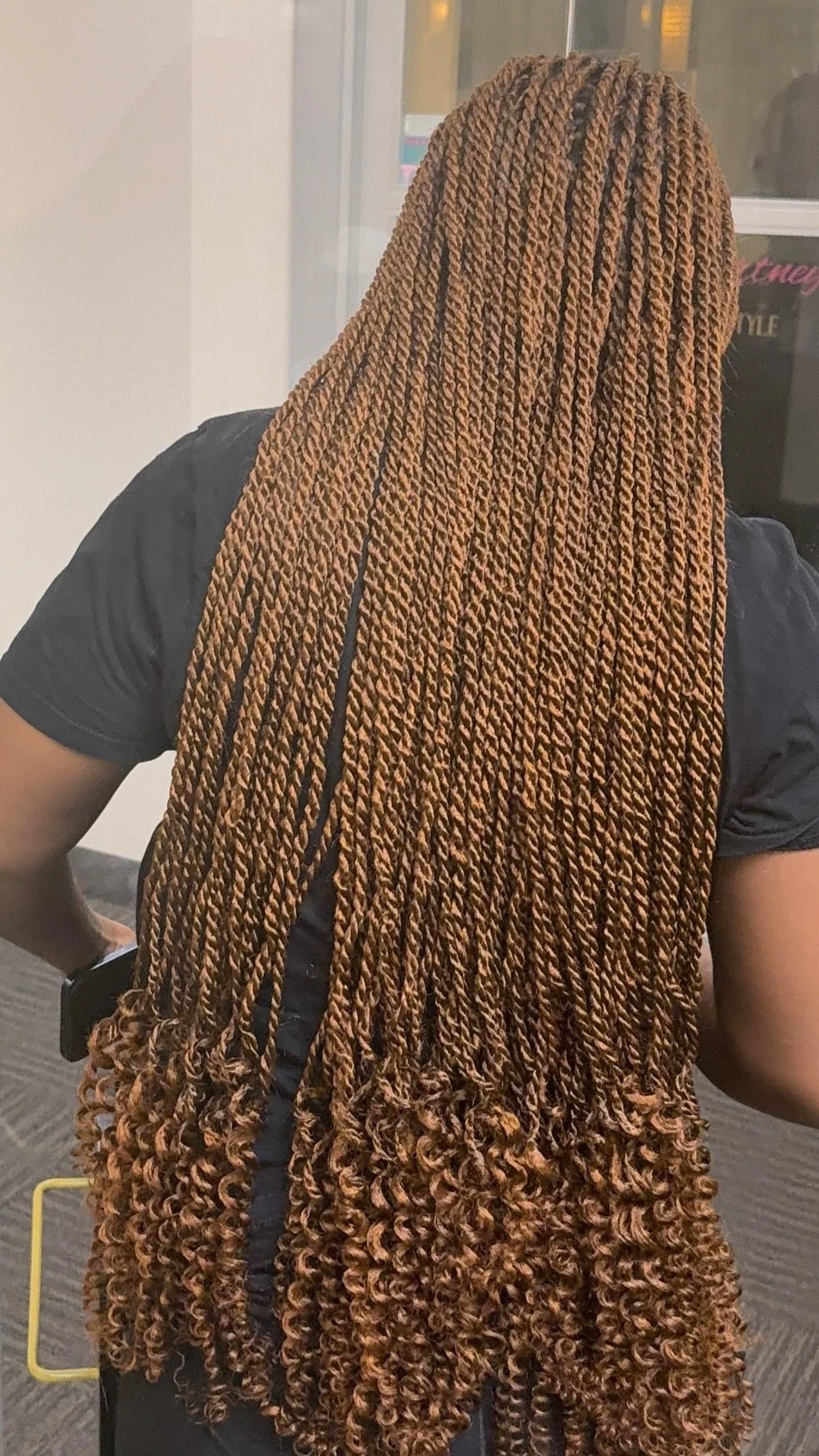 Person with long, brown, twisted faux locs hairstyle, wearing a black T-shirt, standing in front of a window.