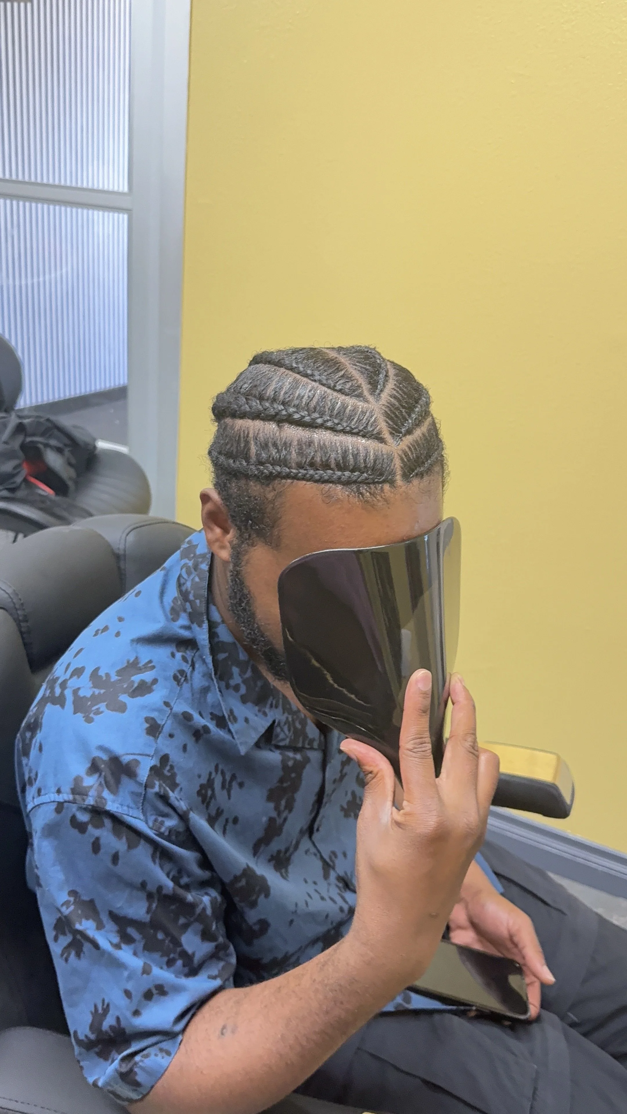 A man with braided hair and a beard sitting in a chair, holding a smartphone and wearing a blue patterned shirt, in a room with yellow and gray walls.