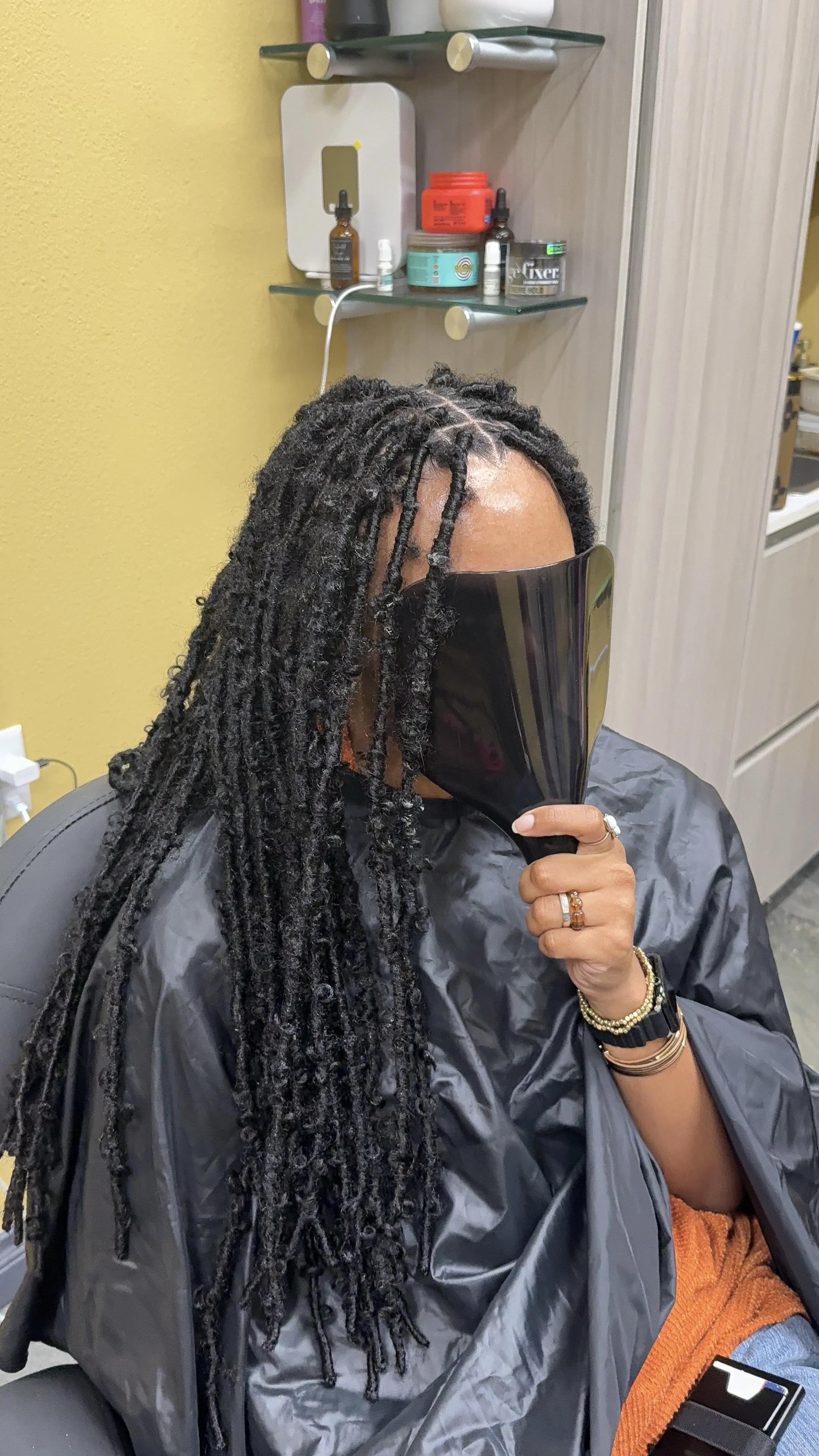 Woman with long dreadlocks sitting in a salon, holding a black reflector in front of her face. She wears a black cape, rings, and bracelets, with a yellow-orange top and blue jeans. Salon shelves with hair products are in the background.