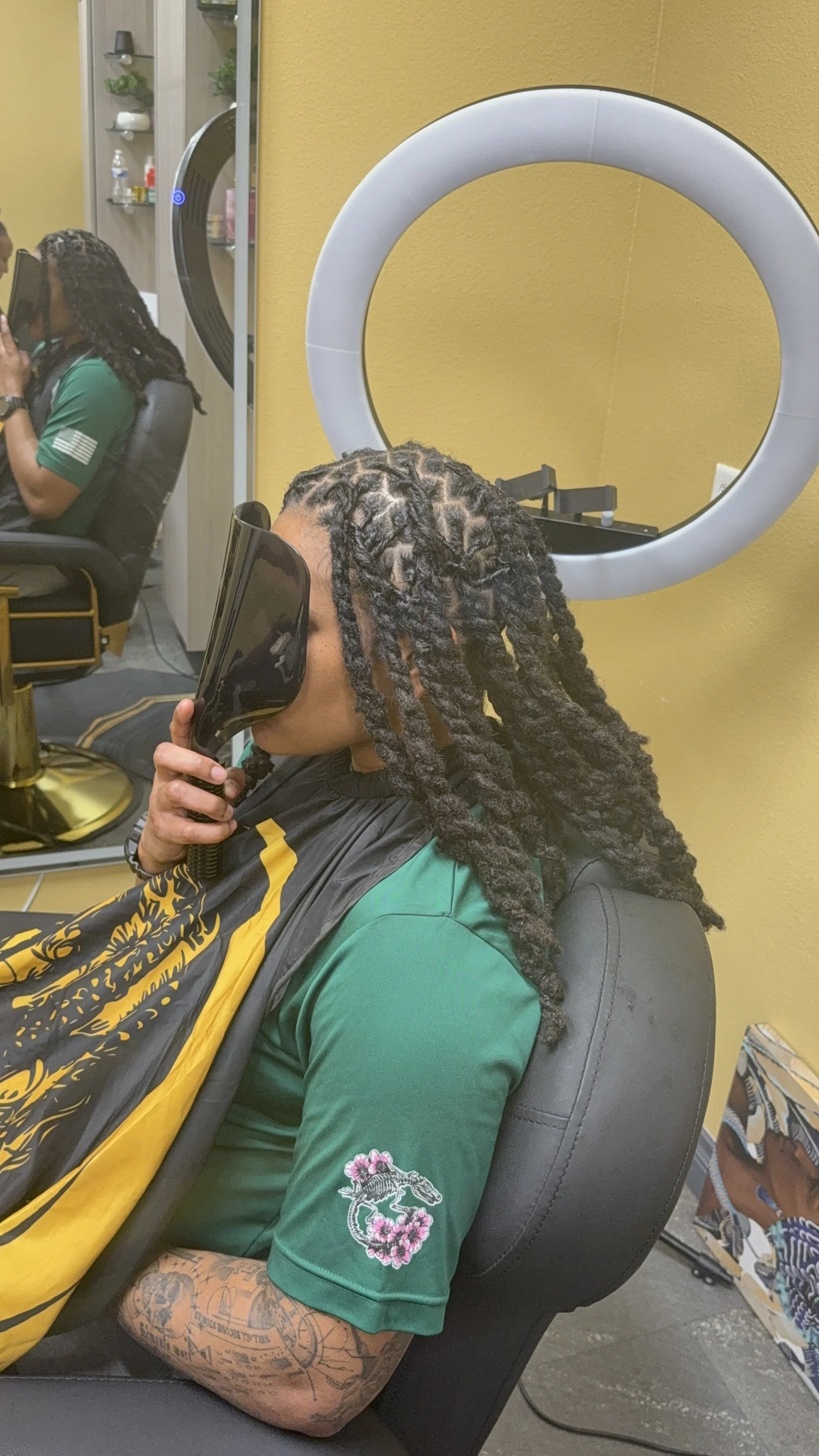 A person with long dreadlocks sitting in a barber's chair, holding a handheld mirror to view their hairstyle, in a salon with a yellow wall and a ring light in the background.