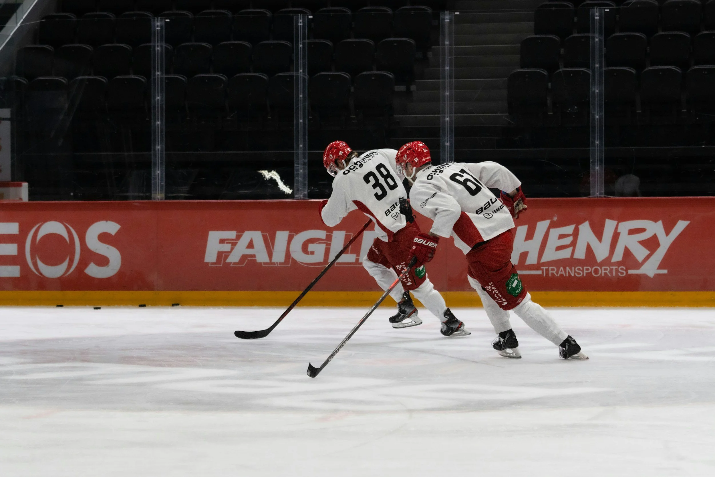 Deux joueurs de hockey en équipement blanc et rouge, puissants, patinent sur la glace lors d'un match, avec des gradins vides en arrière-plan.