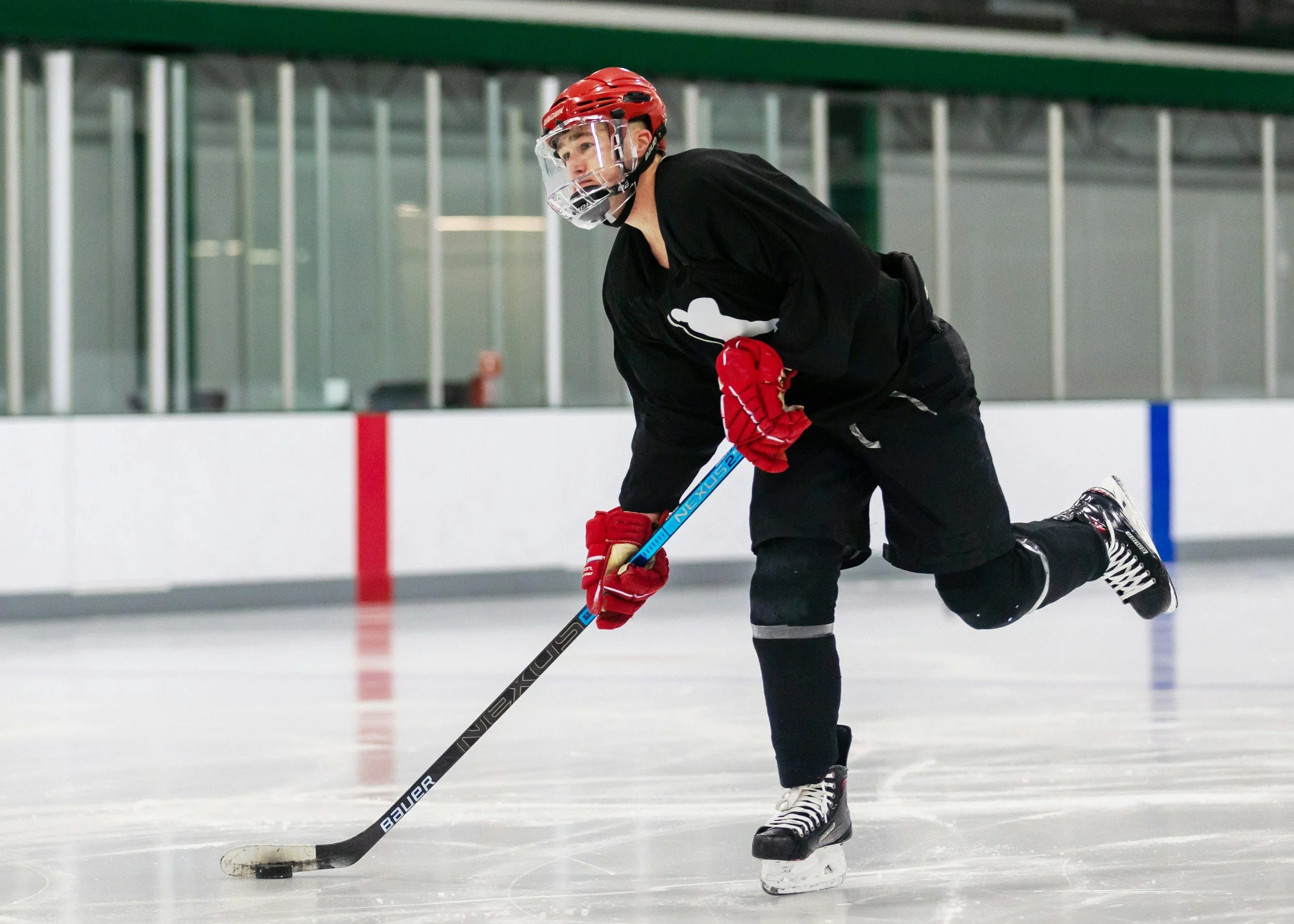 Un joueur de hockey sur glace en action sur la patinoire, portant un casque rouge, une tenue noire, et manipulant une crosse pour jouer avec un palet.