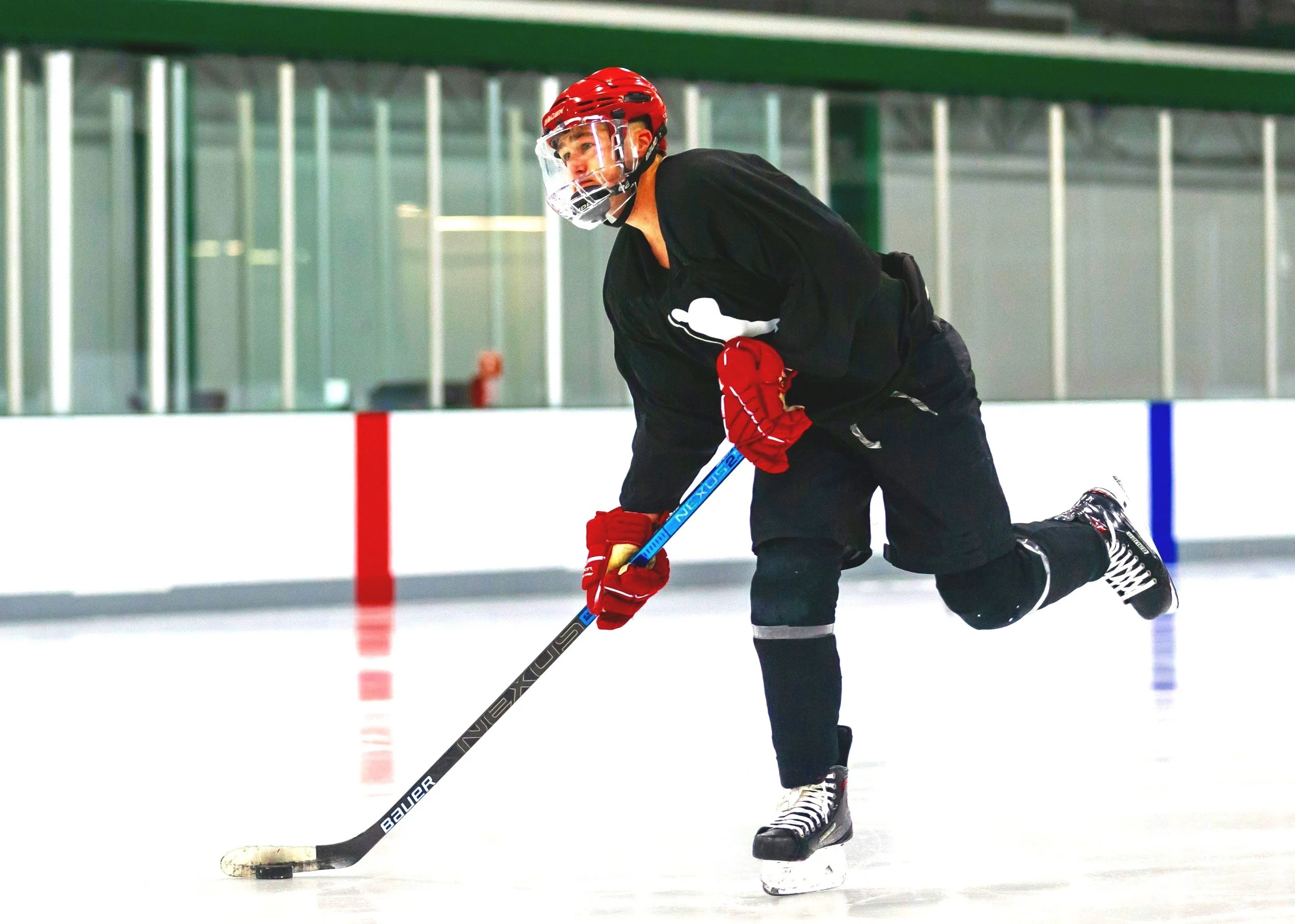 Un joueur de hockey sur glace en position de jeu sur la patinoire, portant un casque rouge, un maillot noir, des gants rouges, et des patins à glace, en train de contrôler la rondelle avec son bâton.