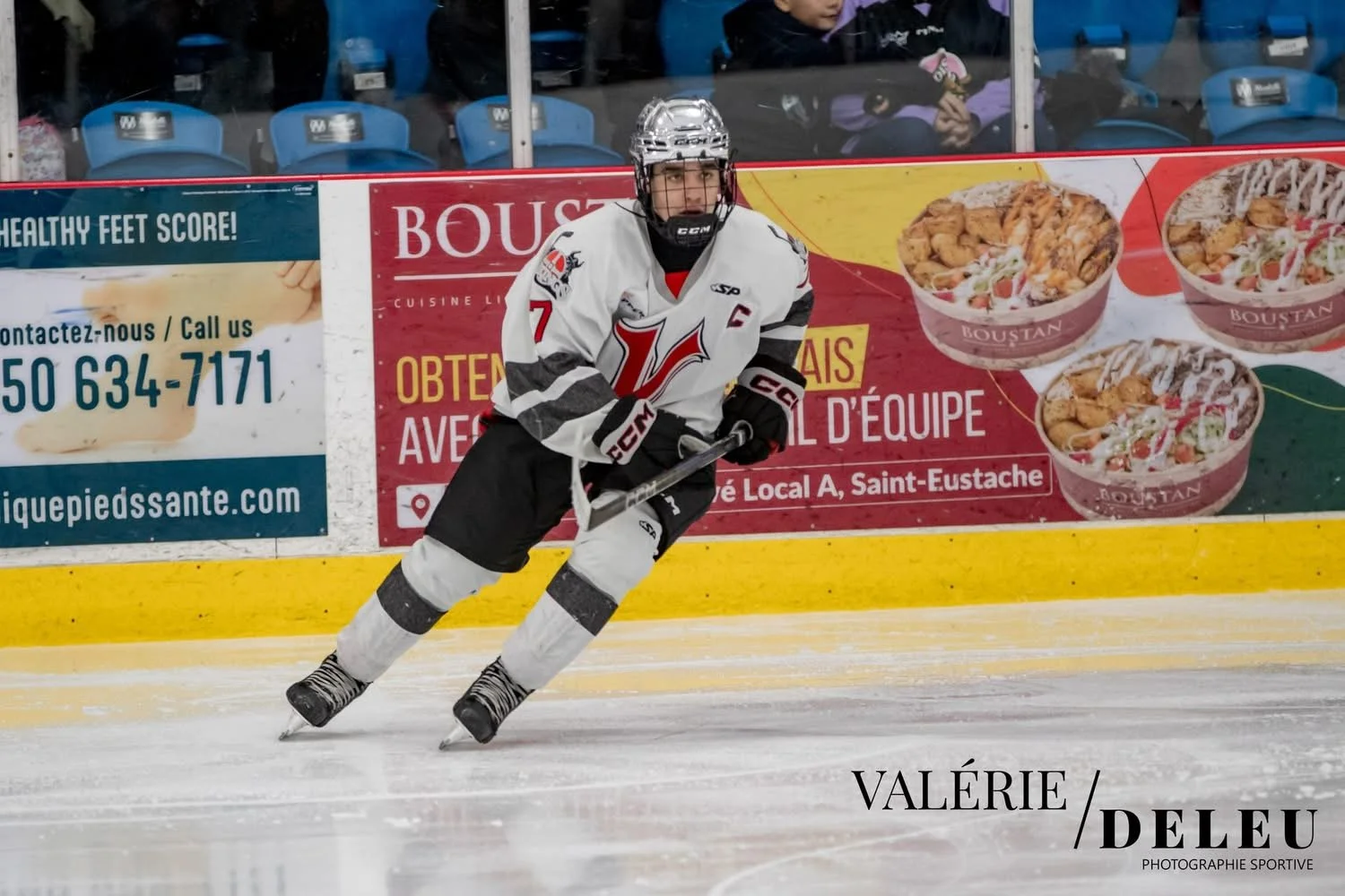 Un joueur de hockey en position sur la glace, portant un uniforme blanc et noir avec le numéro 7 et une C sur la poitrine, en pleine action dans une patinoire vue de face, avec des sièges bleus et des panneaux publicitaires en arrière-plan.