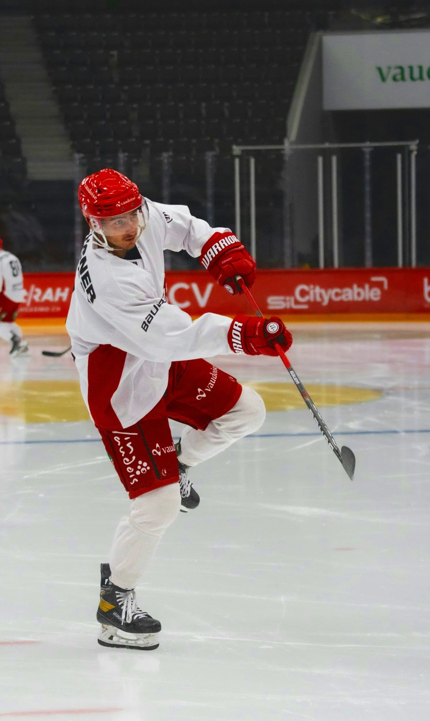 Joueur de hockey sur glace en tenue blanche et rouge, portant un casque rouge, manipulant une crosse de hockey sur la patinoire. Arrière-plan avec des publicités et des gradins vides.
