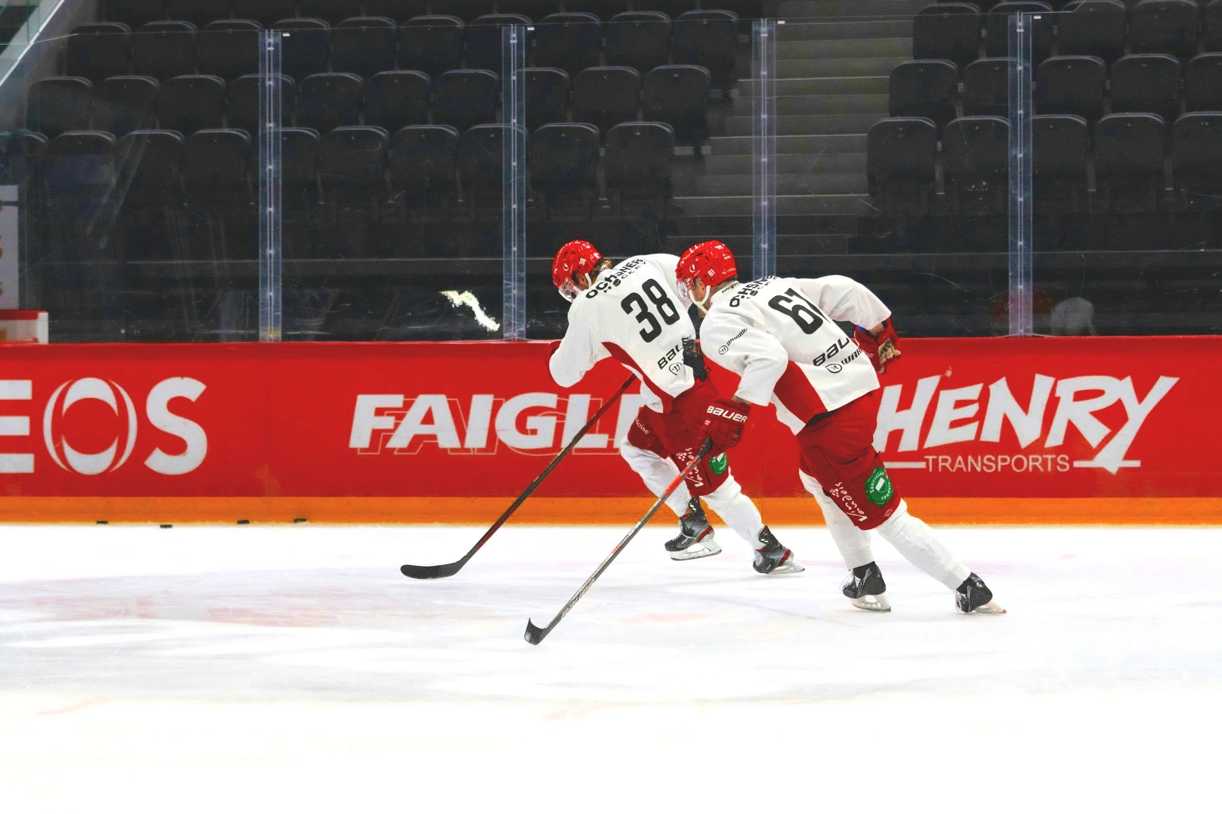Deux joueurs de hockey en train de patiner sur la glace, portant des uniformes blancs avec des numéros 38 et 68, face à une barrière avec des publicités.