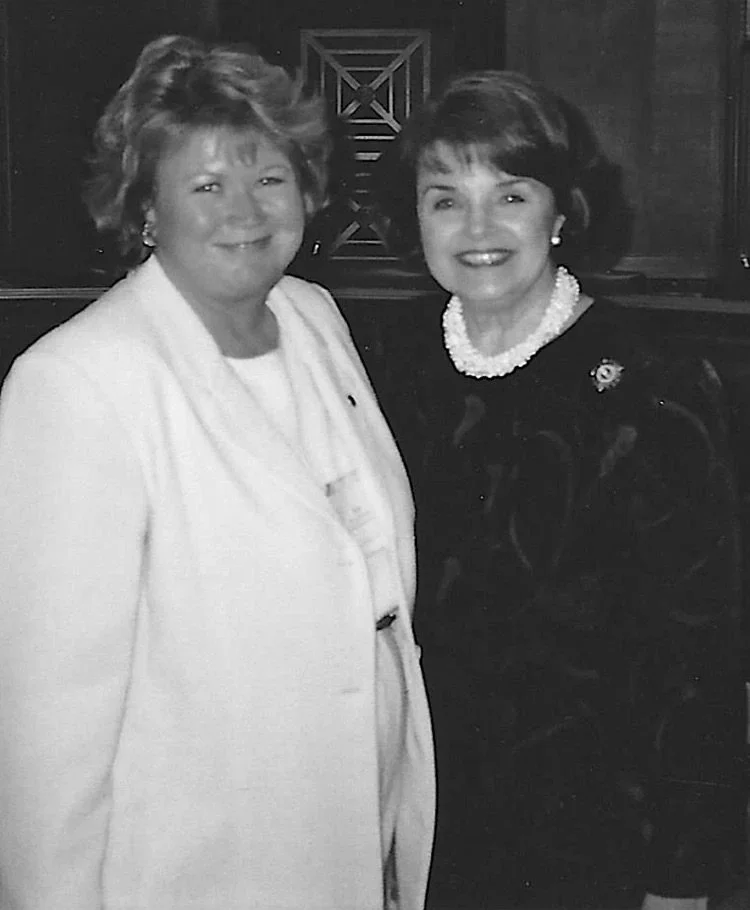 Two women smiling for a black and white photograph; one wearing a white blazer and the other a dark outfit with a pearl necklace.