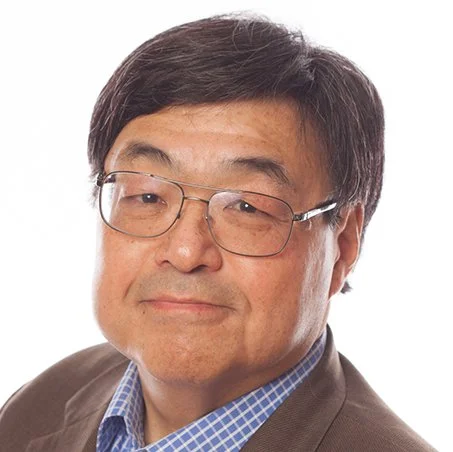 Man wearing glasses, brown jacket, and blue checkered shirt against a white background