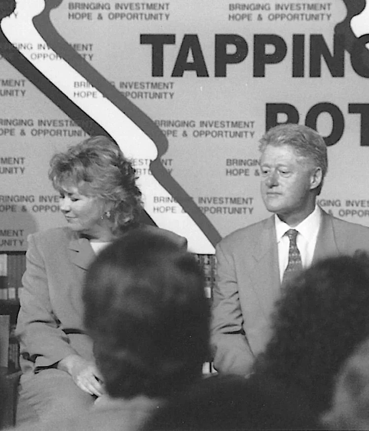A black and white photograph of a man and a woman sitting in front of a backdrop that reads 'TAPPING,' 'BRINGING INVESTMENT HOPE & OPPORTUNITY.' The man is wearing a suit and tie, and the woman is dressed in a blazer. There are people sitting in front of them, partially visible in the foreground.