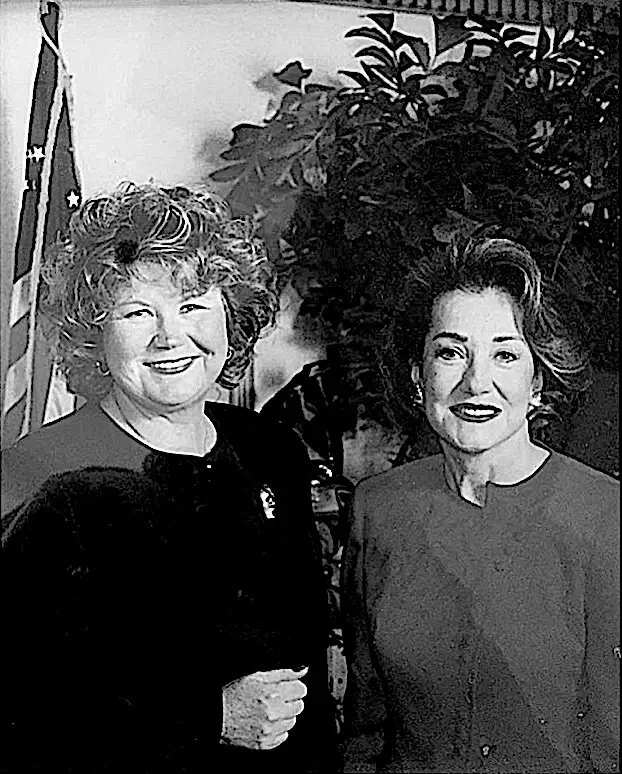 Two women smiling, standing indoors with a U.S. flag and plants in the background.