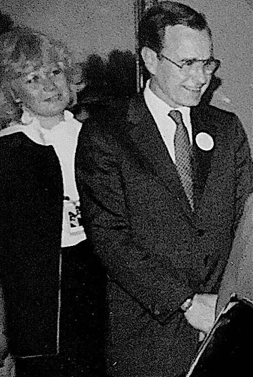 Man in suit with glasses and woman standing behind him