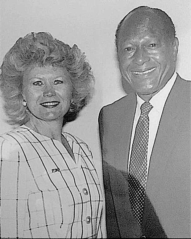 A black and white photograph of a smiling woman and man posing together. The woman is wearing a striped suit jacket and the man is in a suit with a tie.