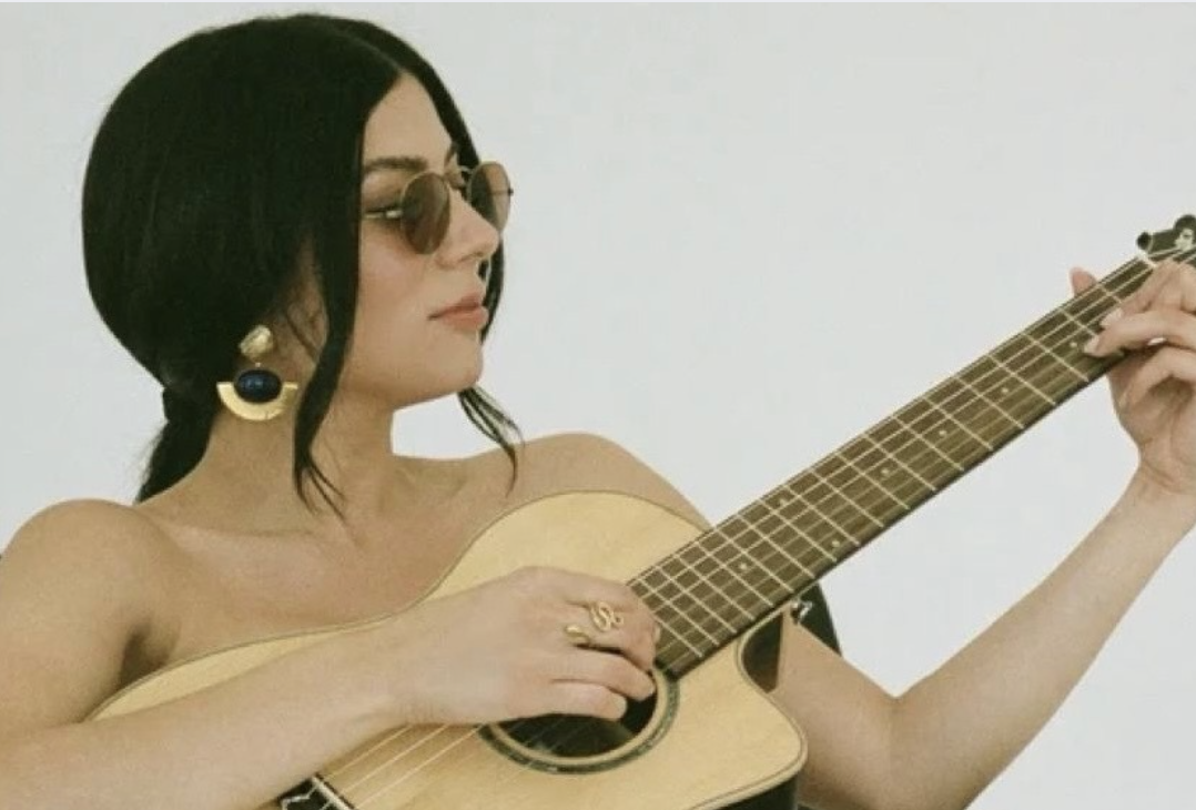 A woman with black hair, wearing sunglasses and earrings, playing an acoustic guitar.