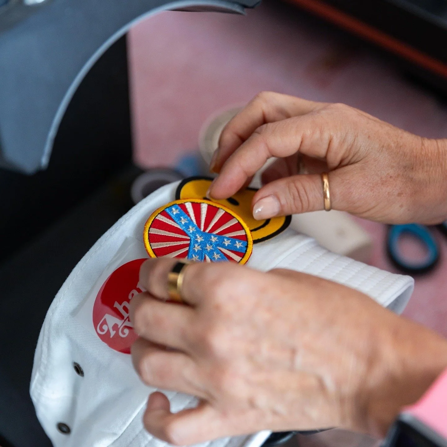 Person placing embroidered patches on a white fabric surface. The patches include a blue peace sign with a red and white striped background and a yellow smiley face. The person is wearing rings on both hands.