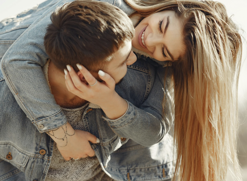 A young couple smiling and cuddling outdoors in sunlight, with the woman embracing the man from behind.