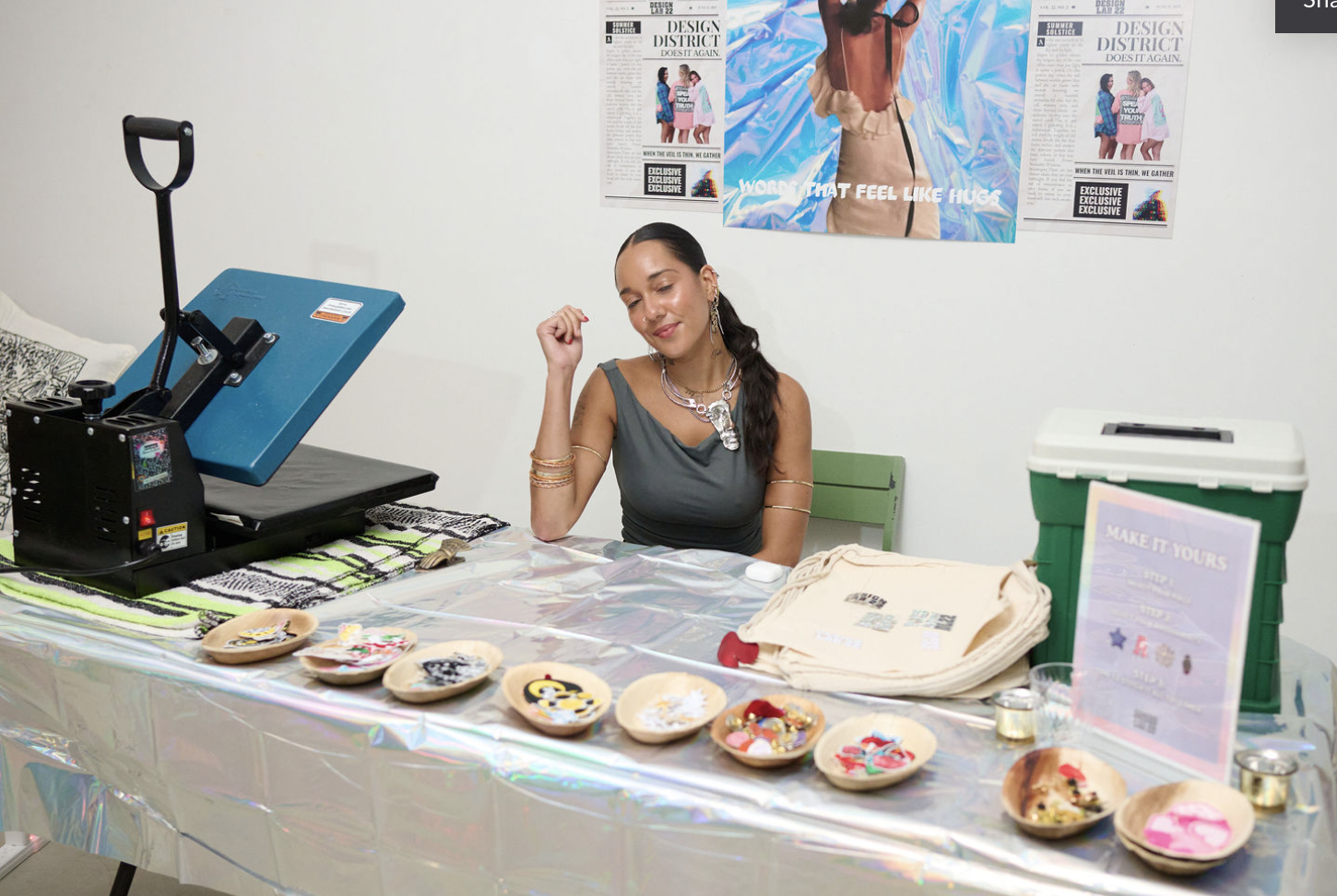 A woman with dark hair and jewelry, sitting at a table with craft supplies, smiling with her eyes closed. The table has various decorated items, small bowls, and a sign that says 'MAKE IT YOURS.' There is a heat press machine and a cooler on the tabl