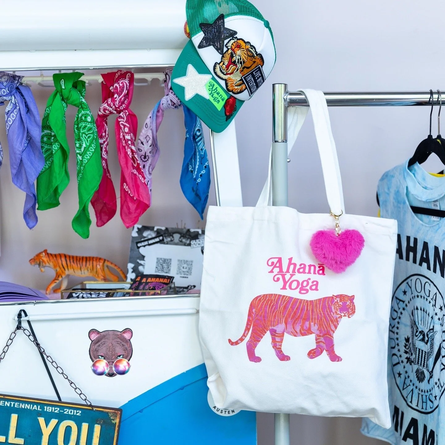 A tote bag with 'Ahana Yoga' and a pink and orange tiger print, hanging on a metal rack among colorful bandanas, hats, and other accessories in a retail display.